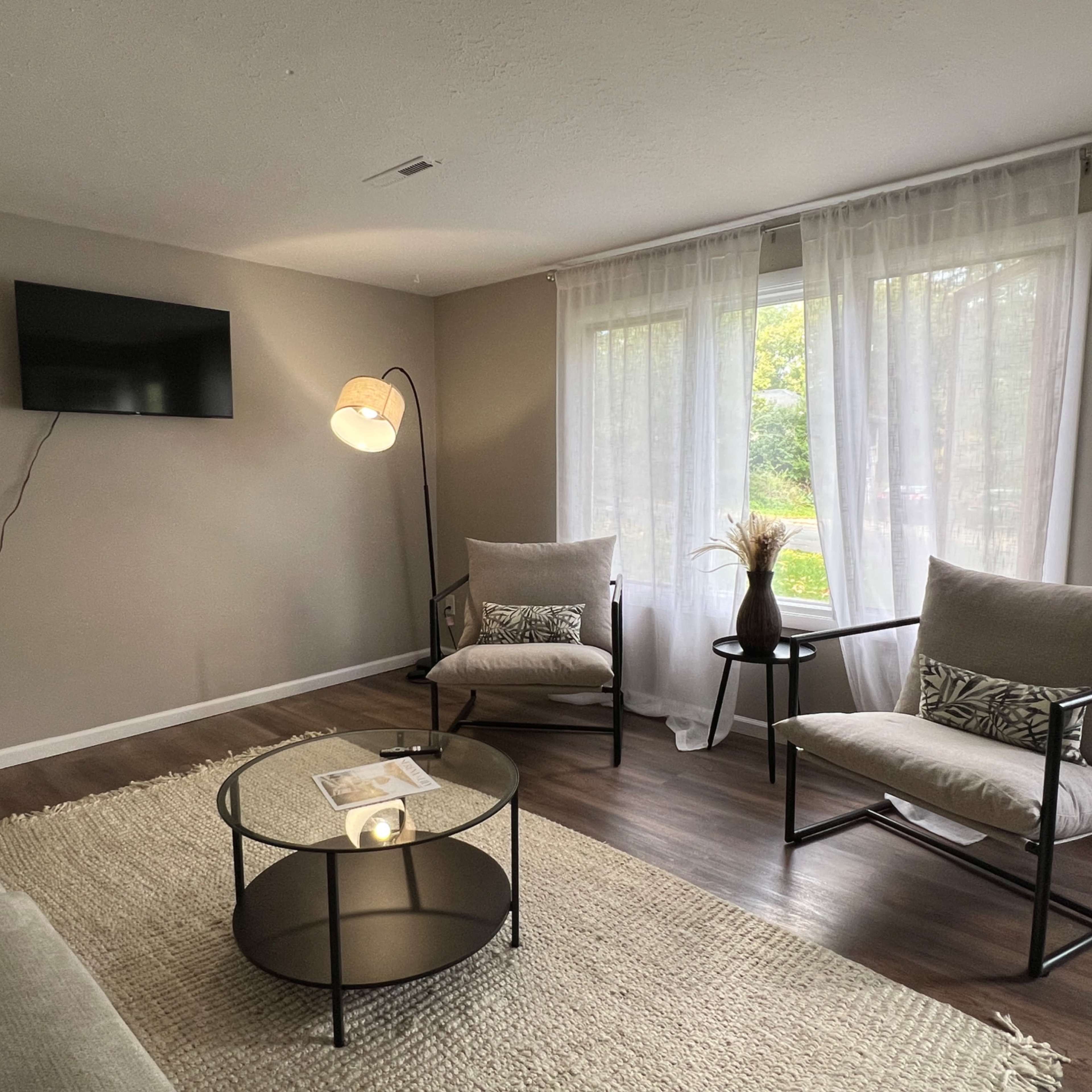 A cozy living room features a television mounted on the wall, two armchairs, a round coffee table, and large windows with sheer curtains allowing natural light.
