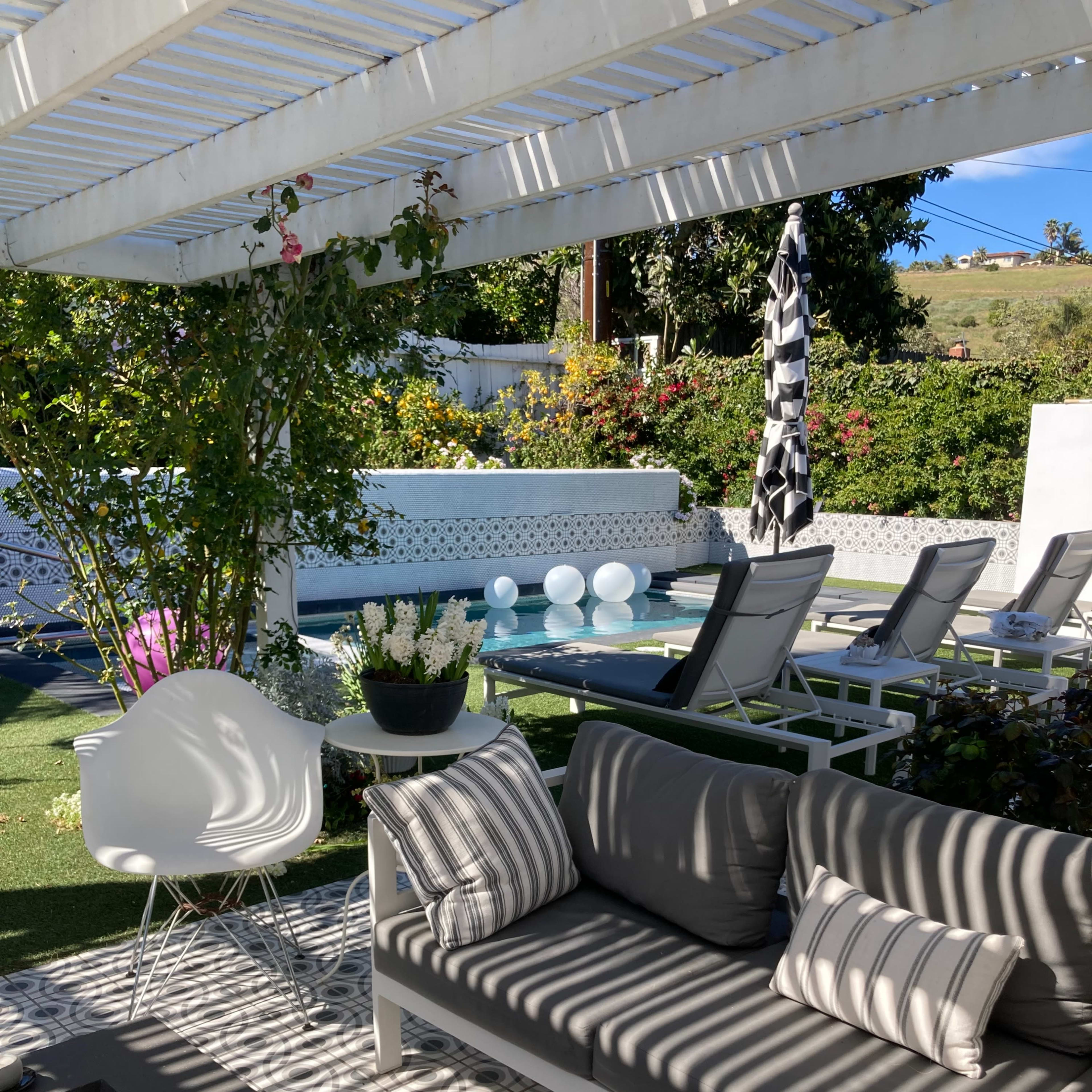 A modern outdoor seating area with a gray sofa, decorative pillows, and a view of a pool surrounded by lounge chairs and trees under a pergola with striped shadows.