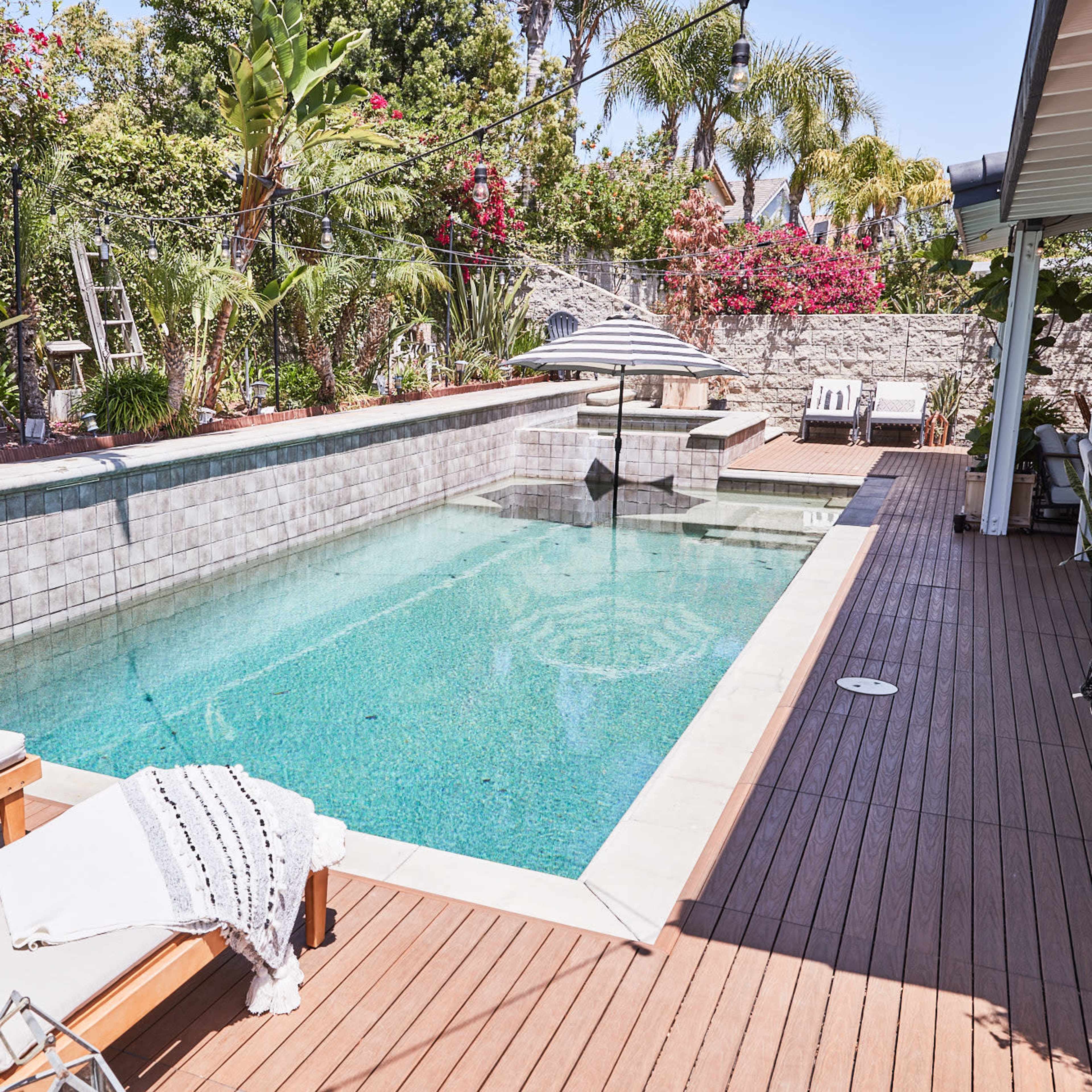 A backyard featuring a pool with clear water, surrounded by tropical plants and a deck with lounge chairs and a table.