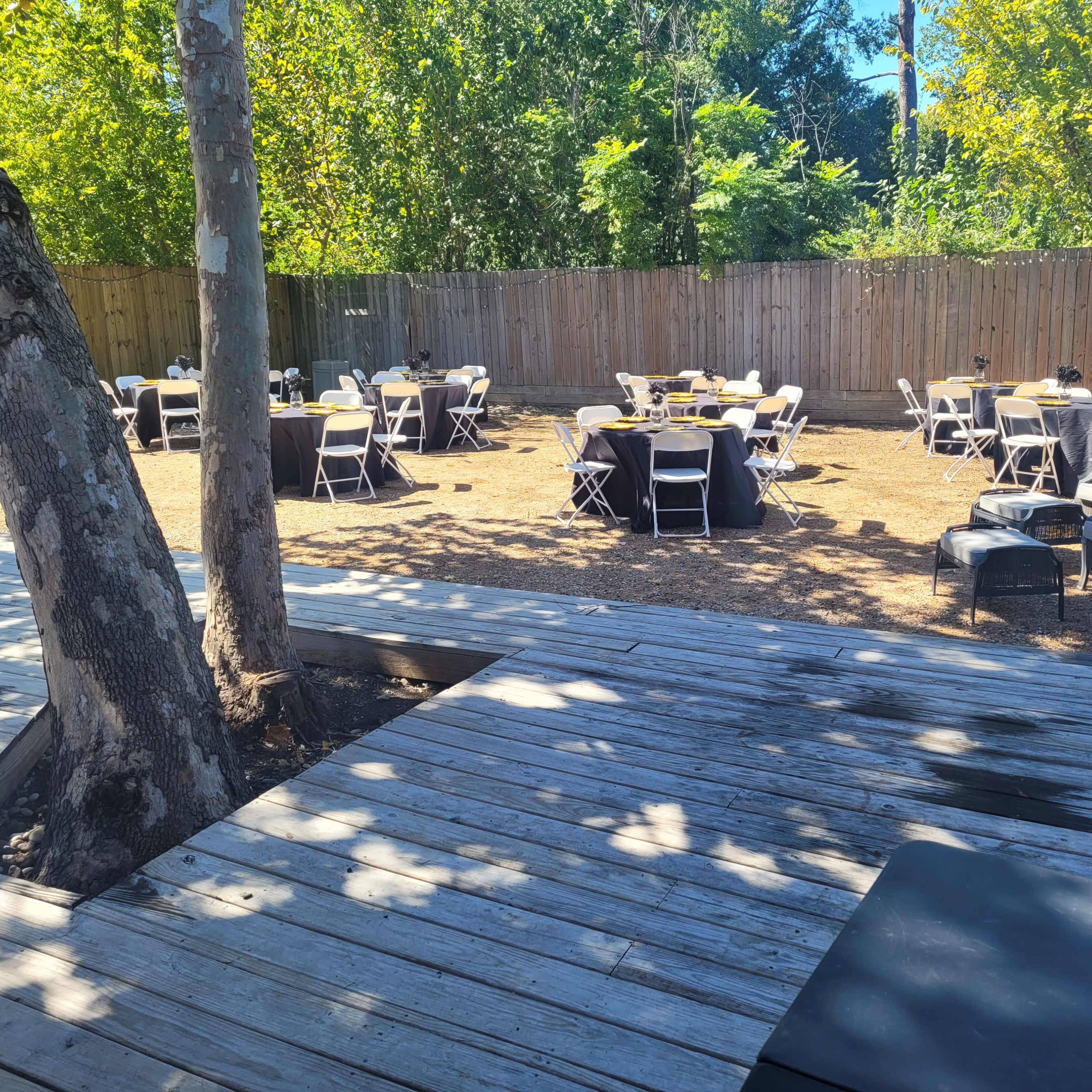 An outdoor event space with several tables set up, surrounded by trees and a wooden fence.