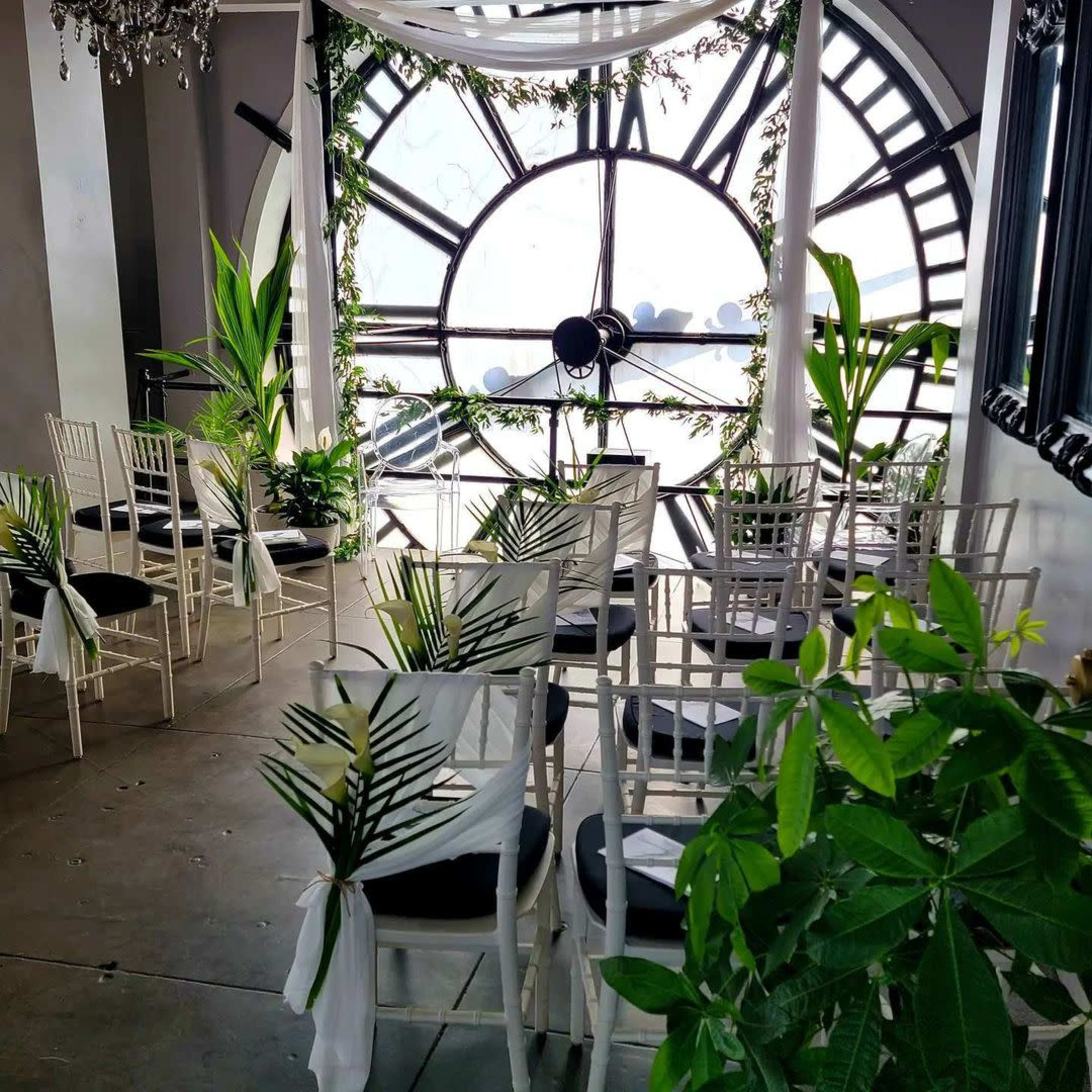 A wedding ceremony setup with white chairs adorned with greenery, arranged in front of a large clock window.
