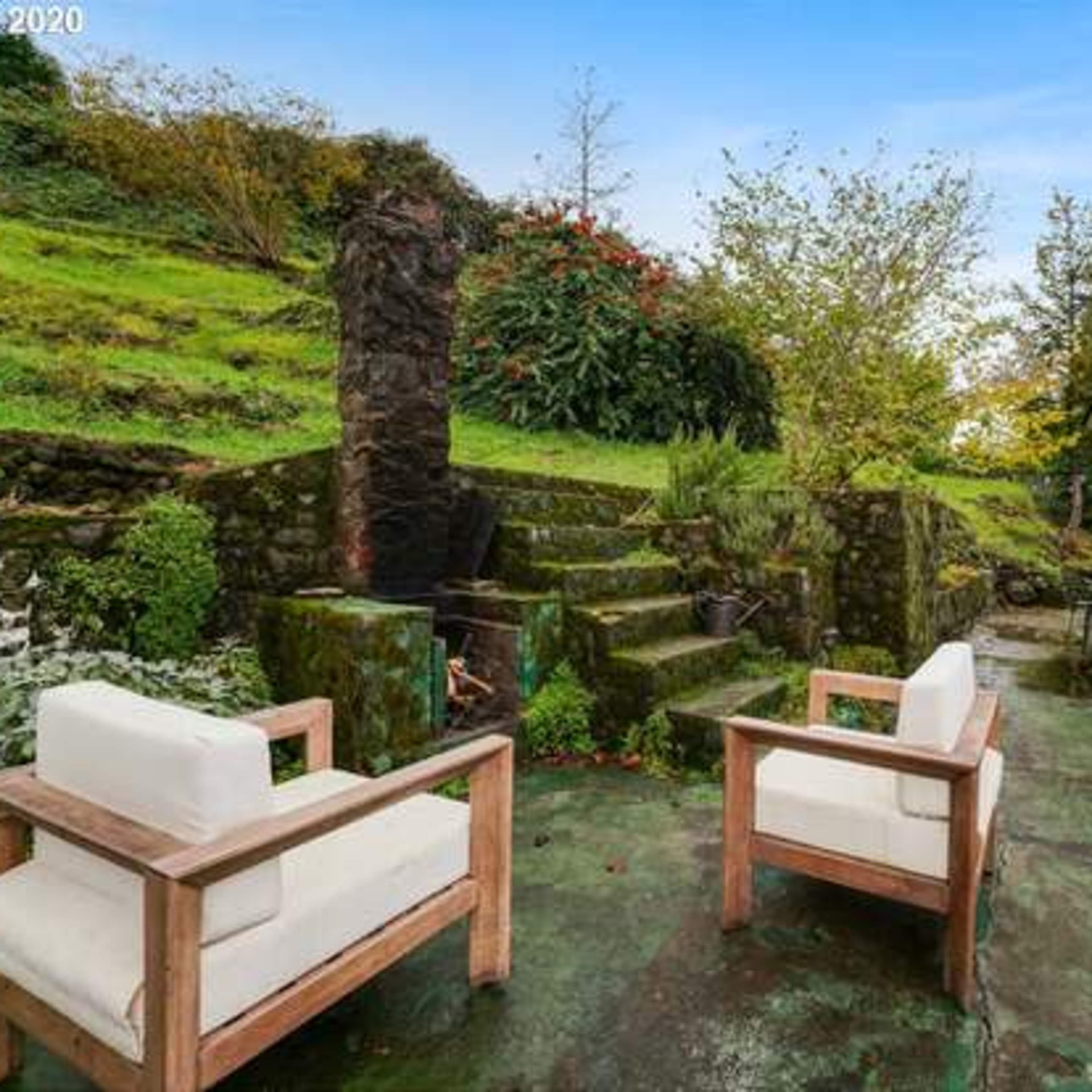 An outdoor patio area with two wooden chairs, steps leading up to a grassy hillside, and a small building with a white door.