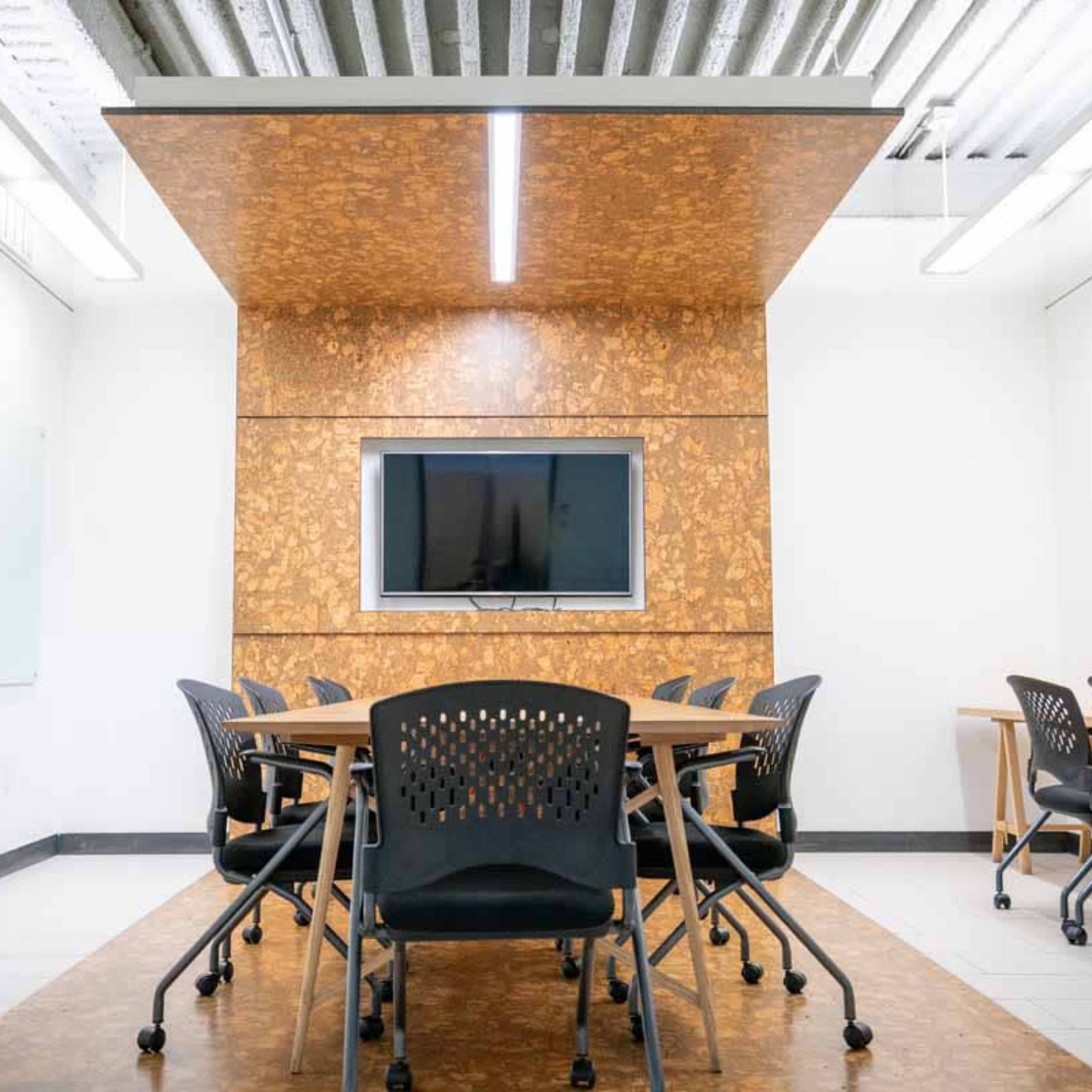 A modern conference room features a central table surrounded by black chairs, with a wall-mounted television and cork paneling in the background.