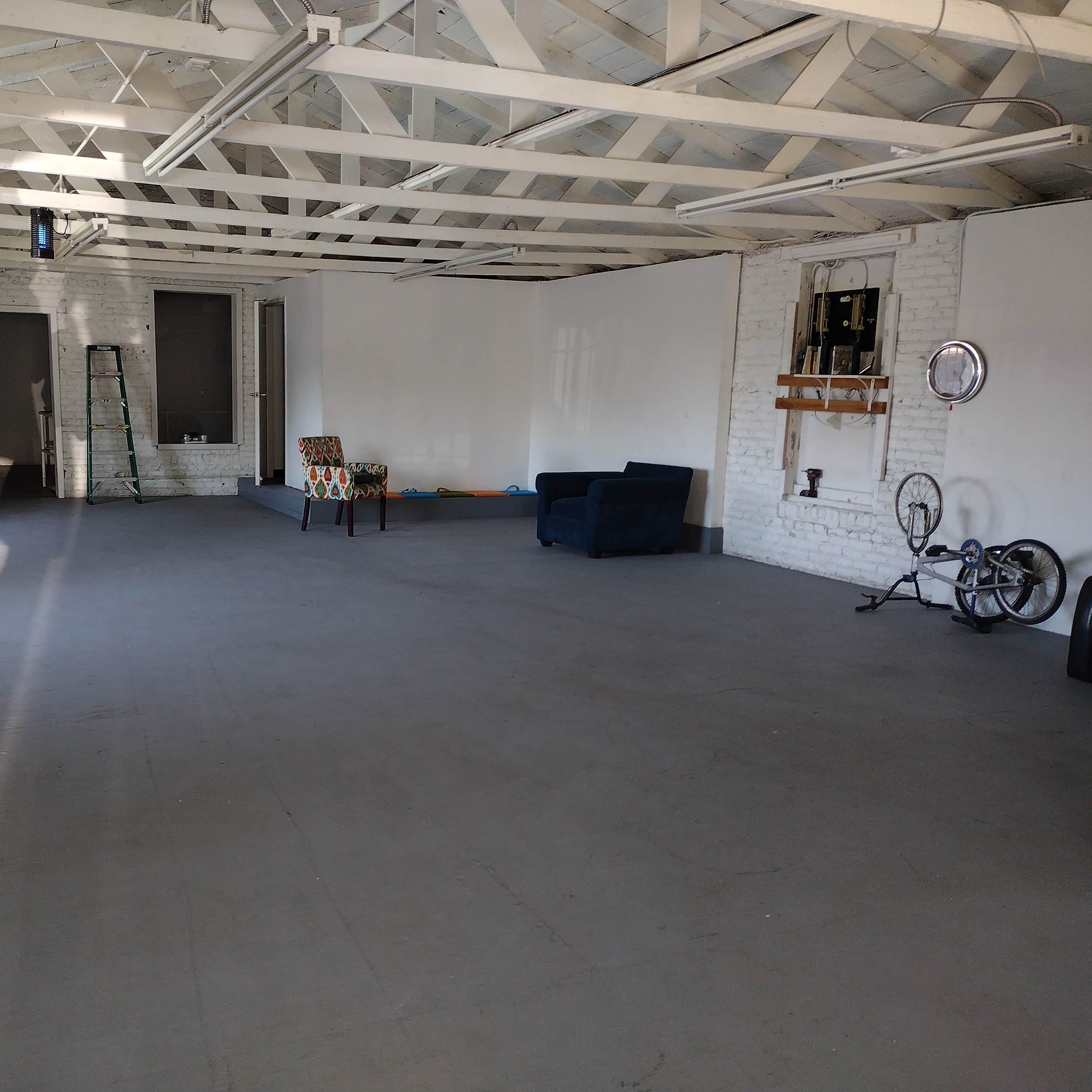A spacious, empty room with a white brick wall, wooden beams, and minimal furniture including a blue couch and a patterned chair.