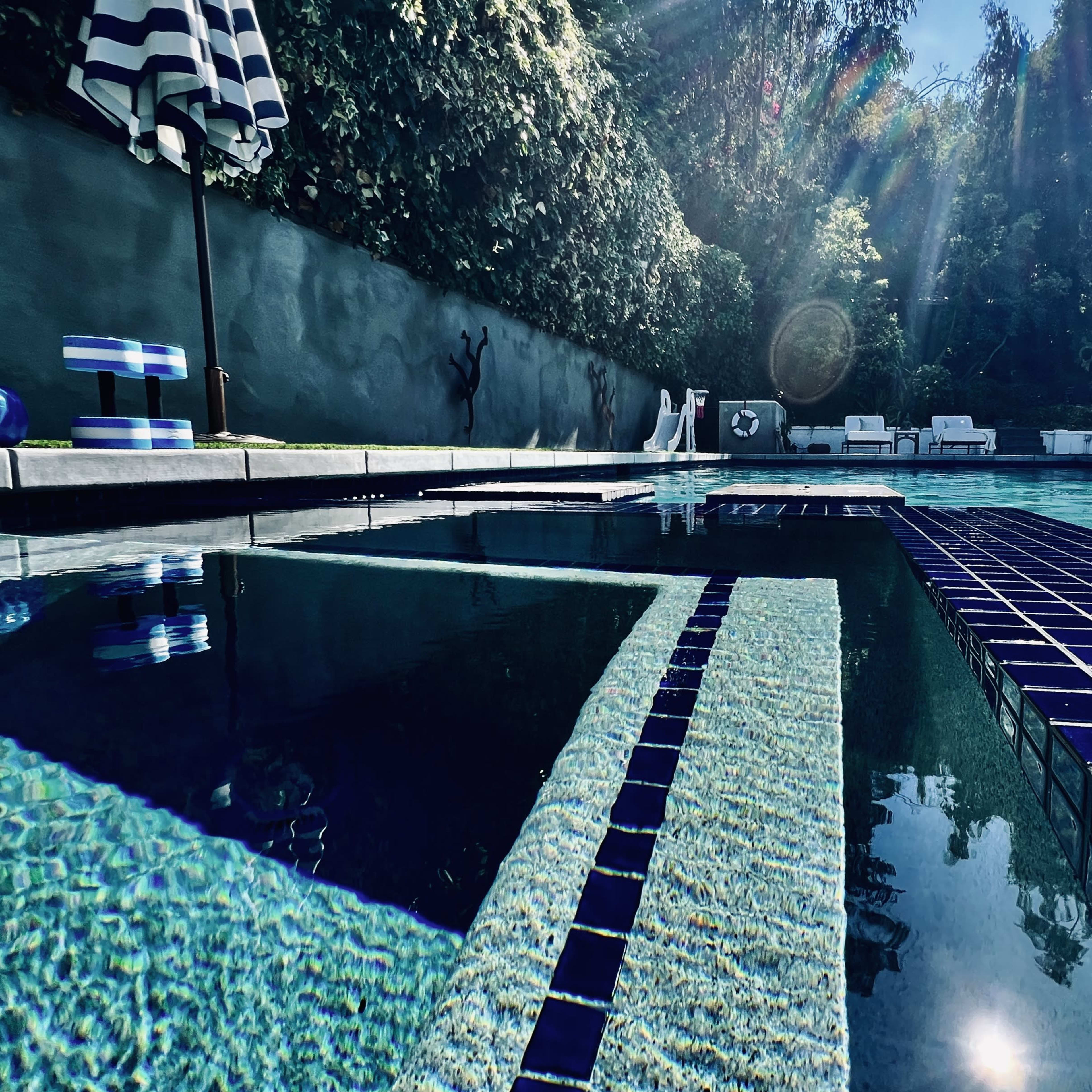 A swimming pool with blue tiles, surrounded by foliage and lounge chairs under a bright sky.