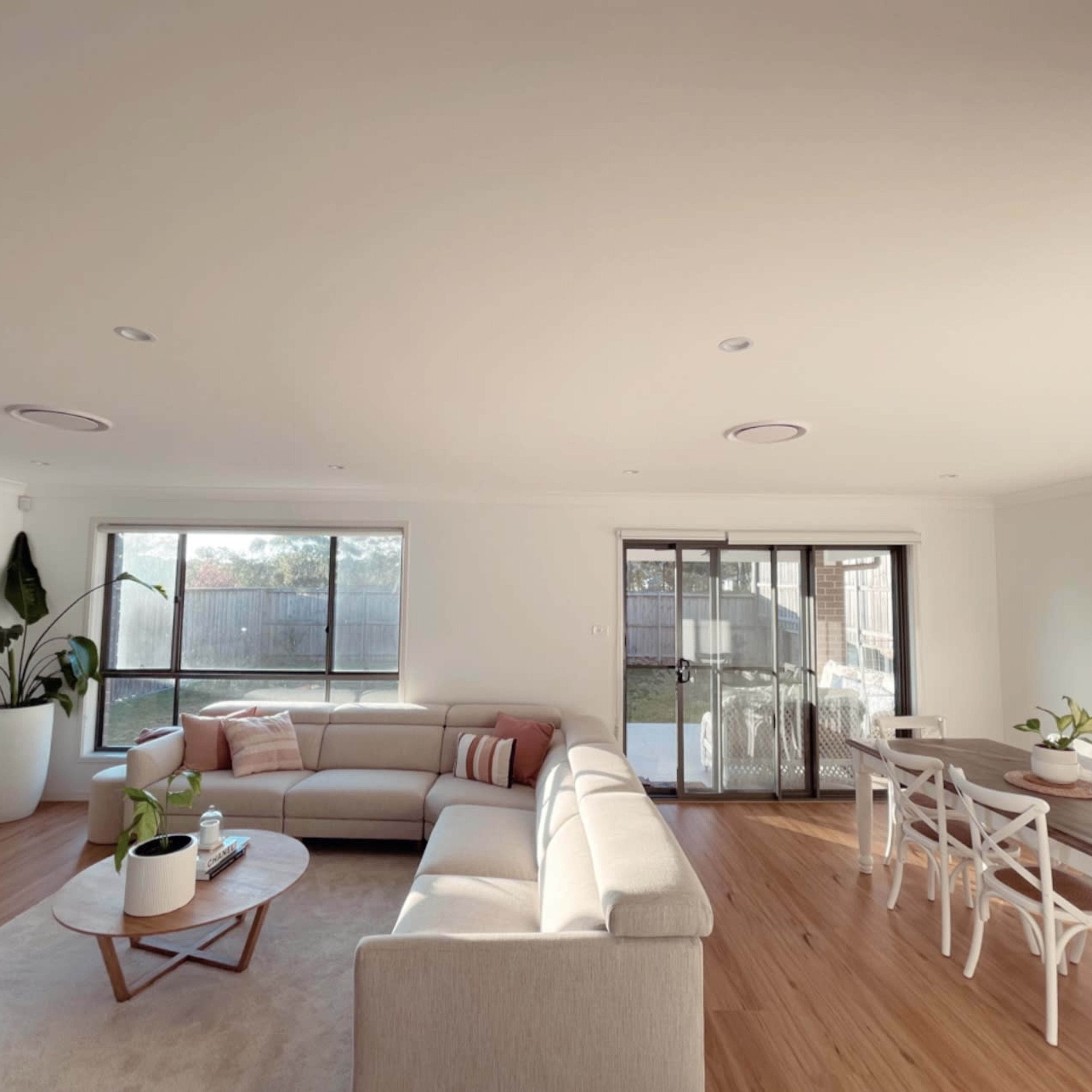 A spacious living and dining area with a light-colored sectional sofa, a circular coffee table, a dining table with chairs, and large windows overlooking an outdoor space.
