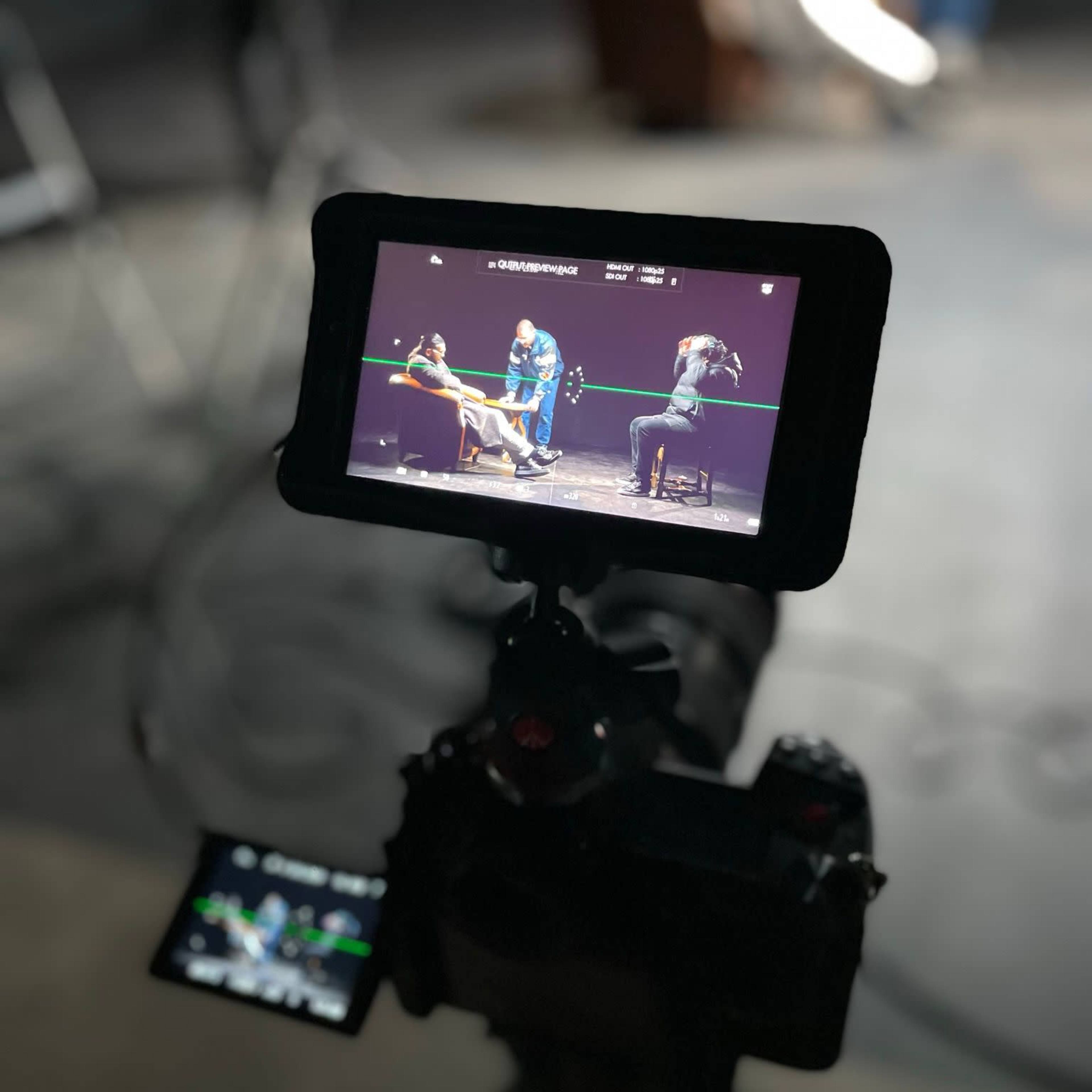 A camera on a tripod is focused on two people seated on stage, captured on a video monitor in a studio setting.
