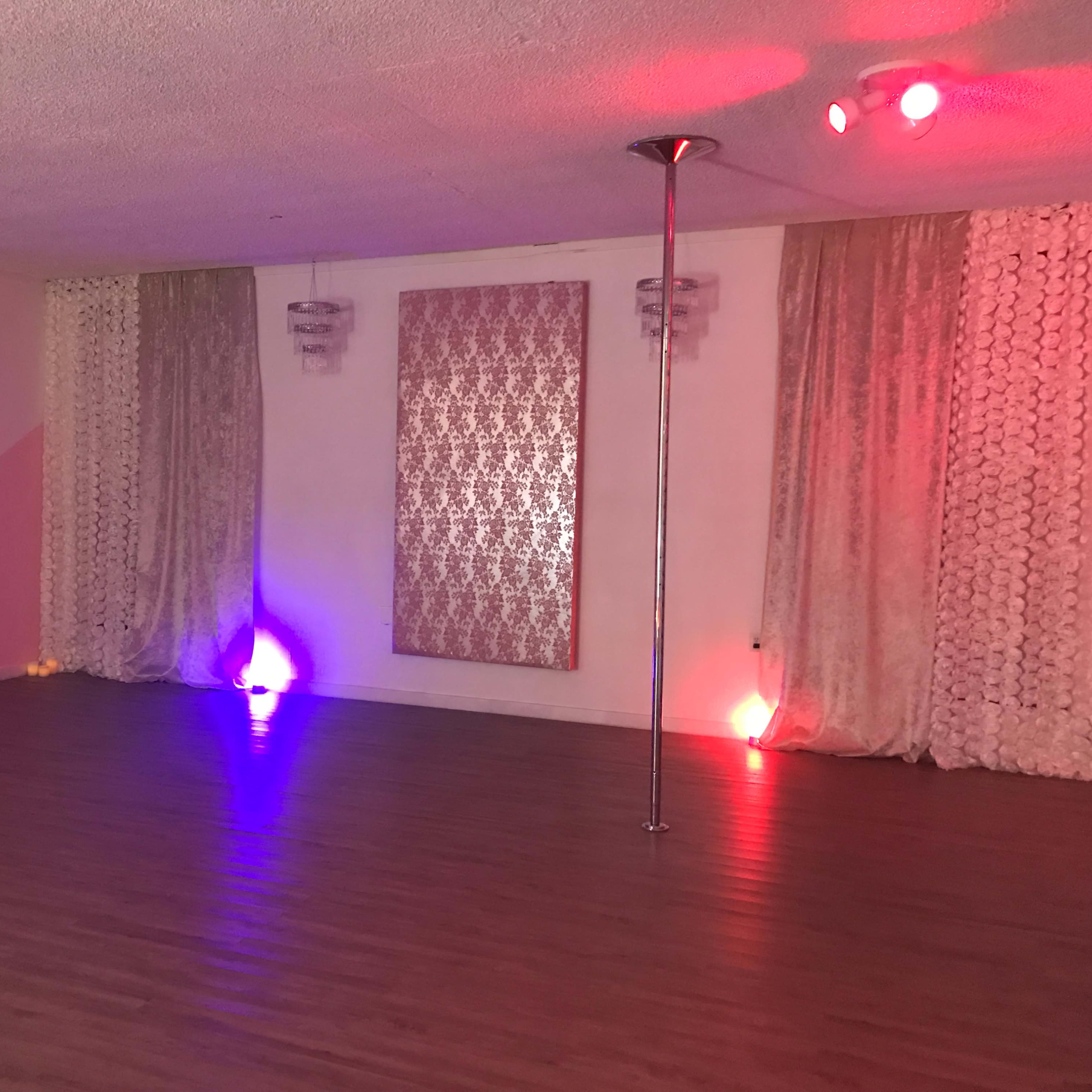 A dance studio with a polished wooden floor, a vertical pole in the center, and ambient lighting from colored lamps along the walls.
