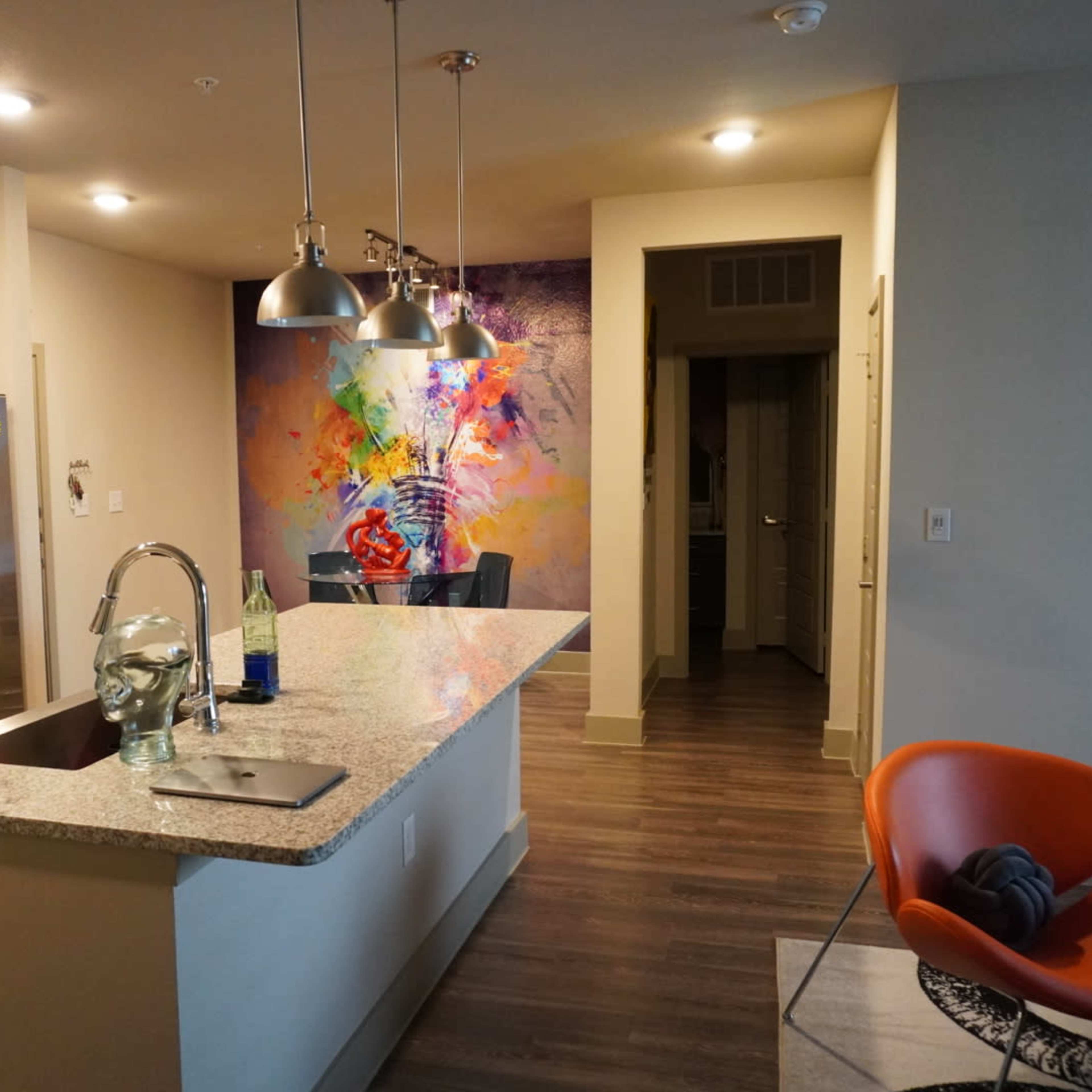 A modern kitchen with granite countertops, stainless steel appliances, and a vibrant mural on the wall, leading to a hallway with an orange chair nearby.