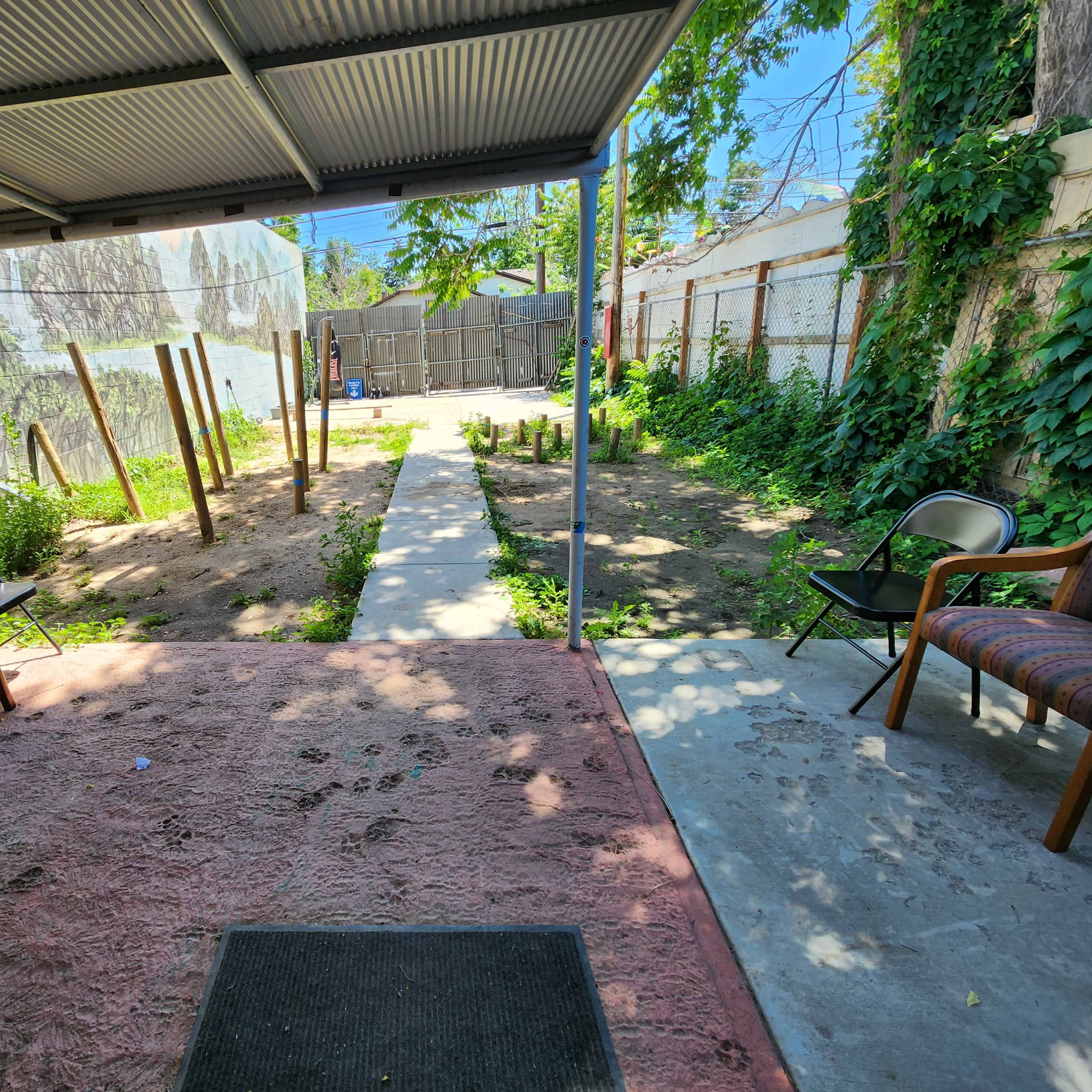 A shaded outdoor area with several empty chairs and a dirt pathway leading to a garden space with wooden poles and greenery in the background.
