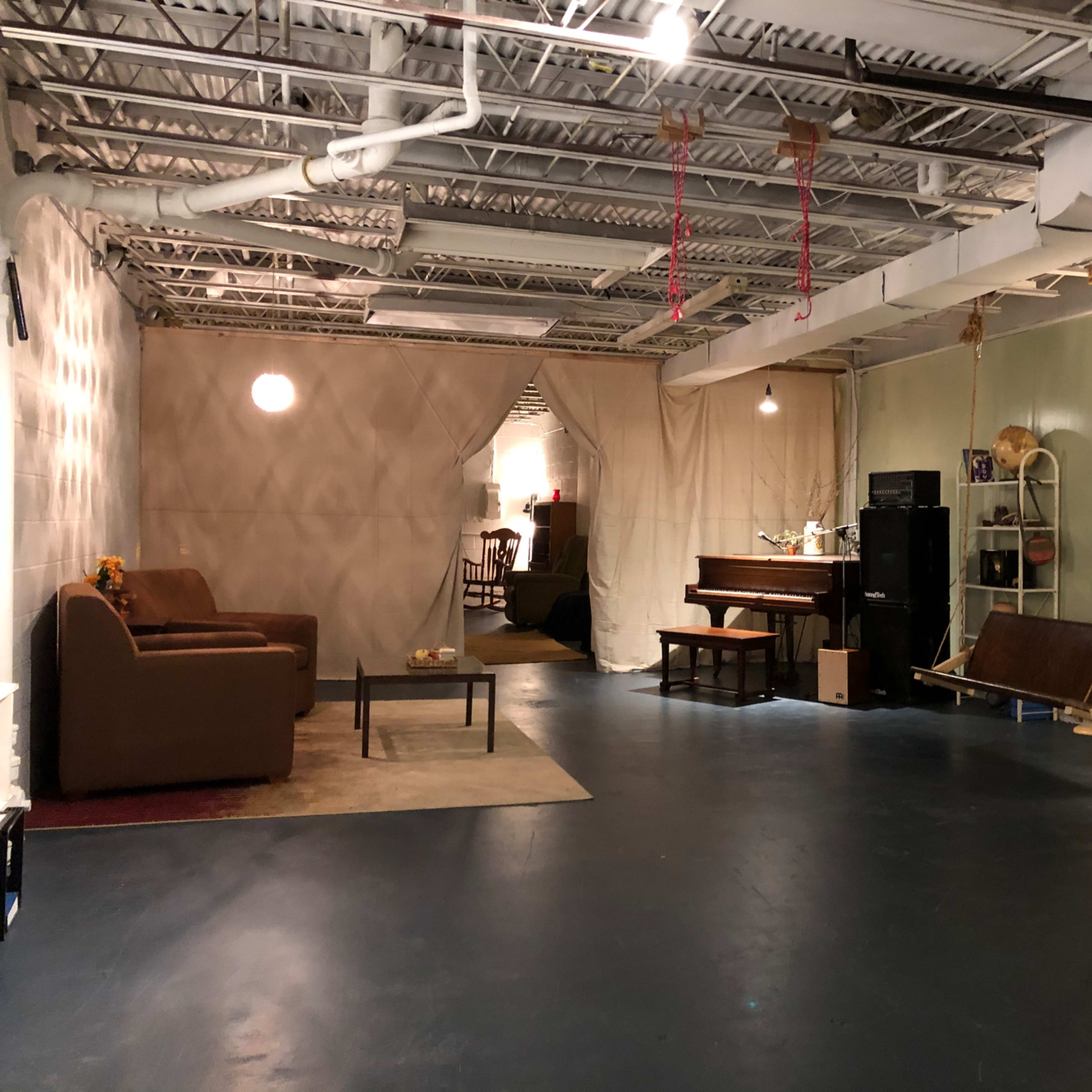 The interior space features a living area with two brown couches, a piano, a swing chair, and a small table with a laptop, all under a ceiling with exposed pipes.