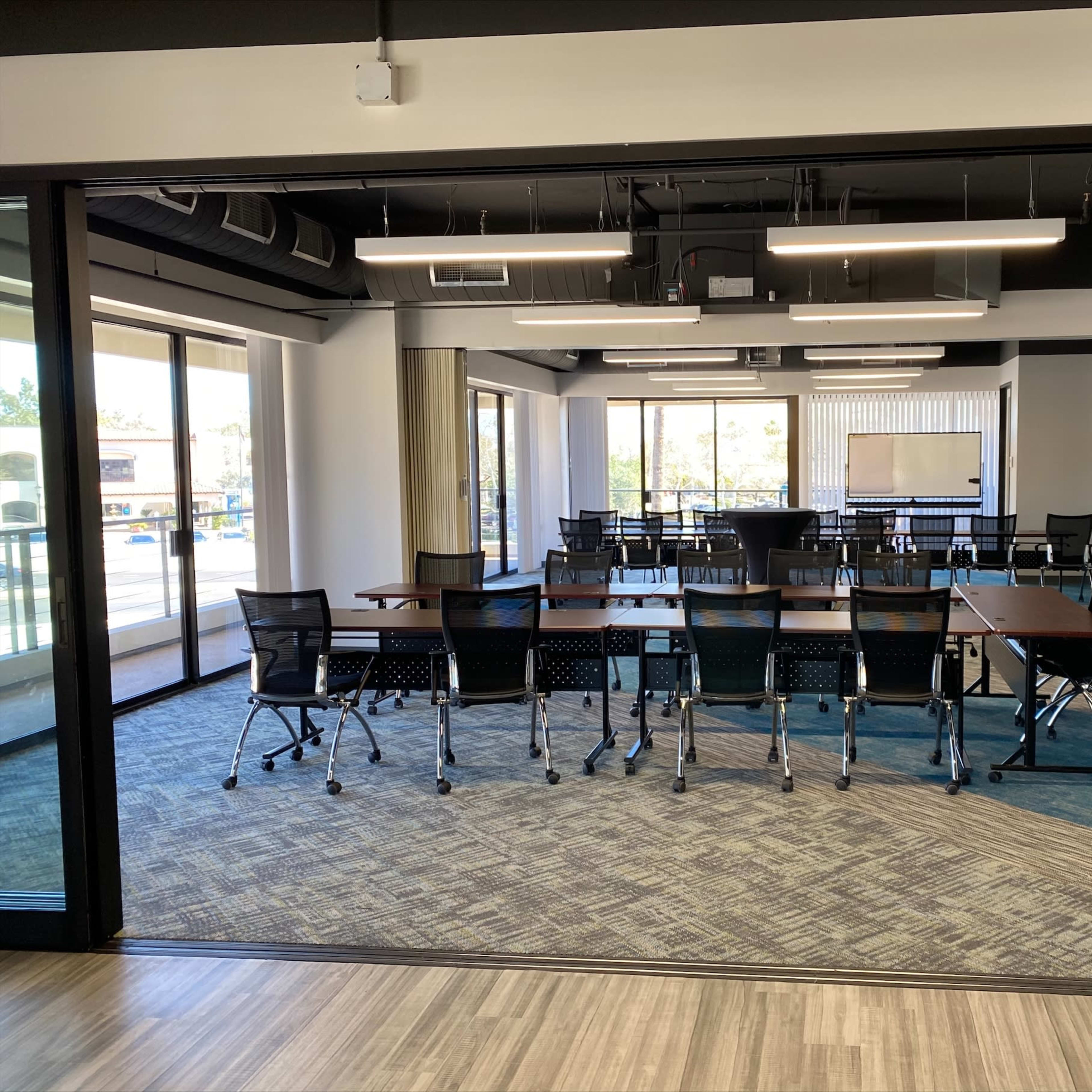 A conference room is set up with multiple tables and chairs, featuring large windows that provide natural light and views of the outdoors.