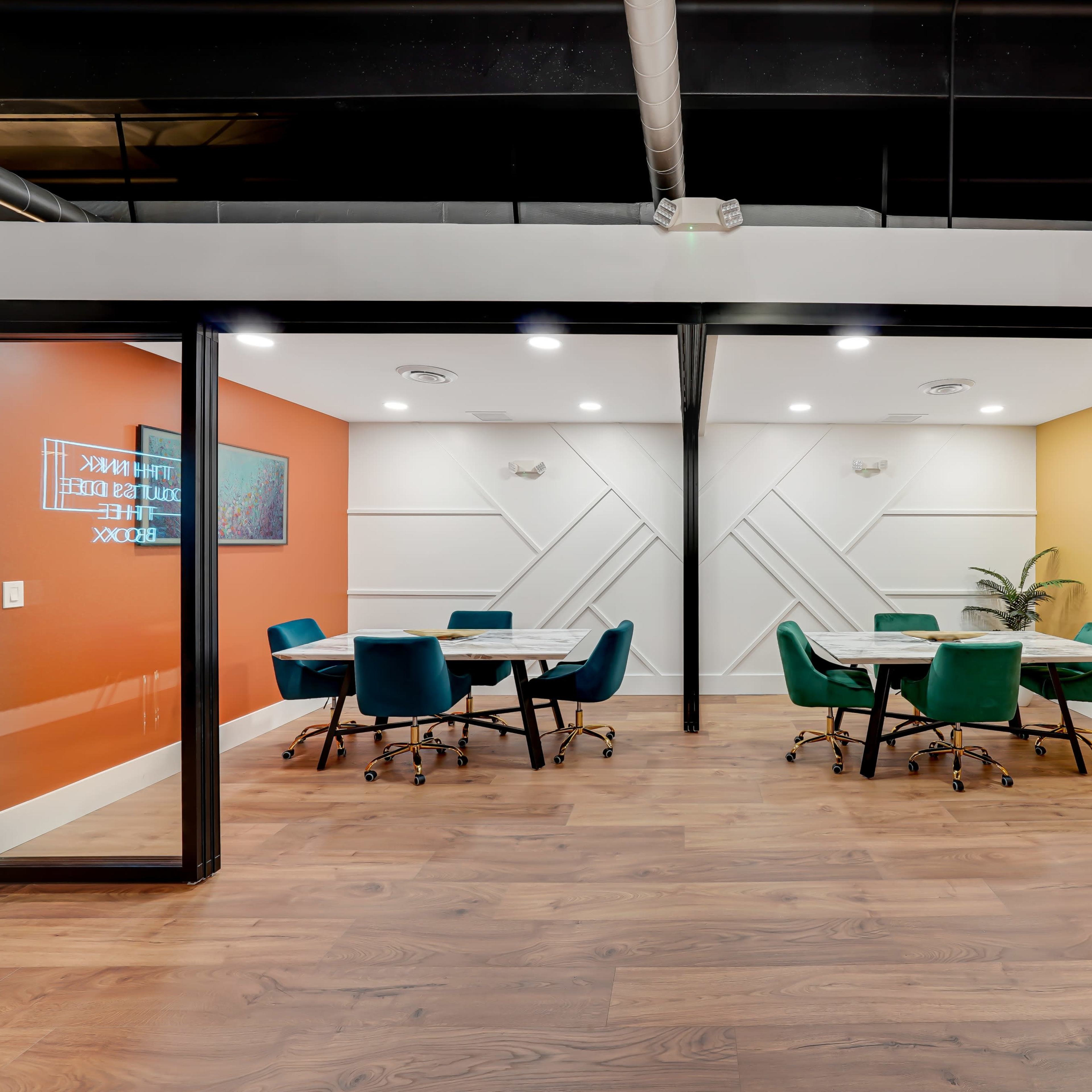 Two meeting rooms with glass walls, each containing a round table surrounded by teal chairs, set against orange and yellow accent walls.