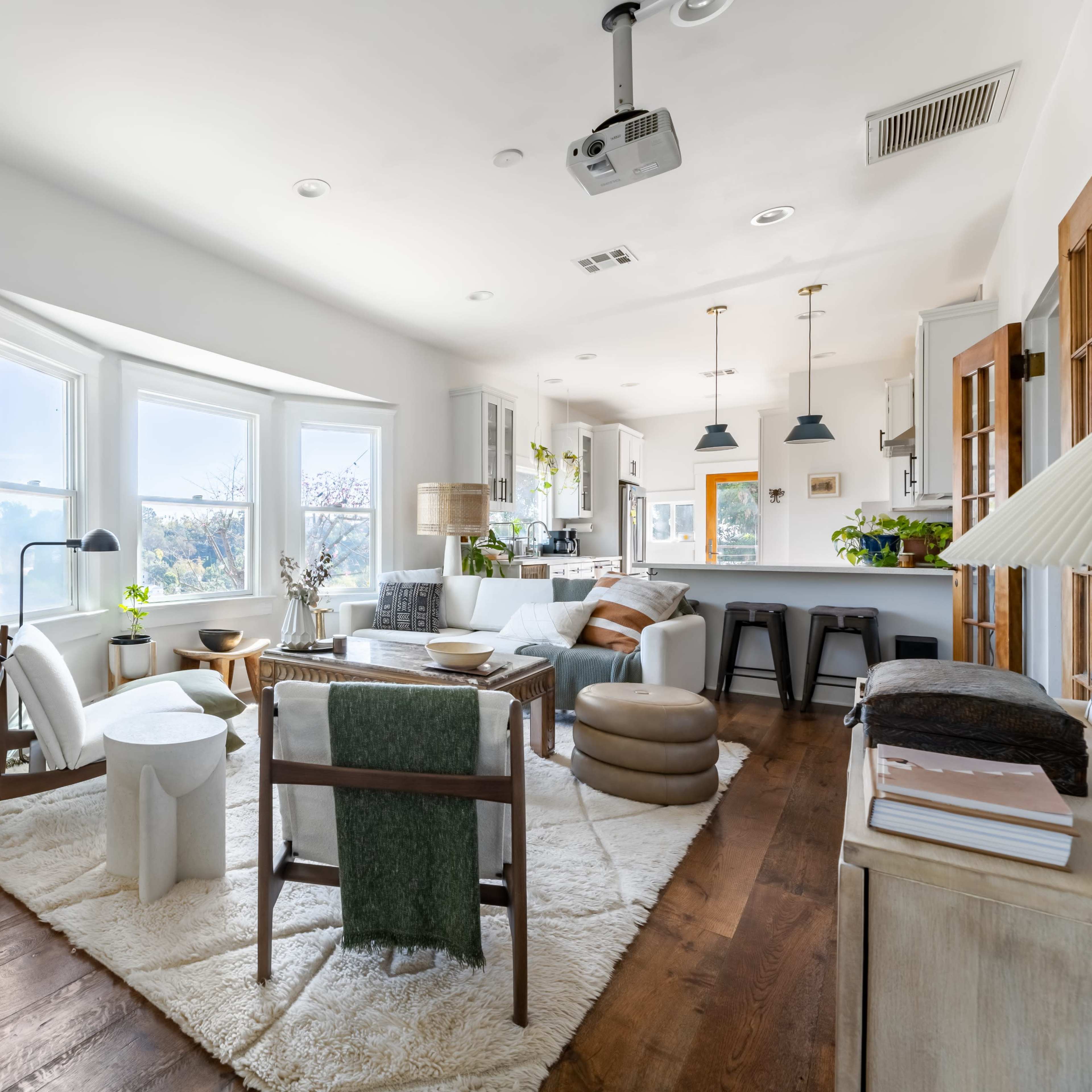 A modern living room with large windows, a light-colored rug, and various seating options, connected to a kitchen area.