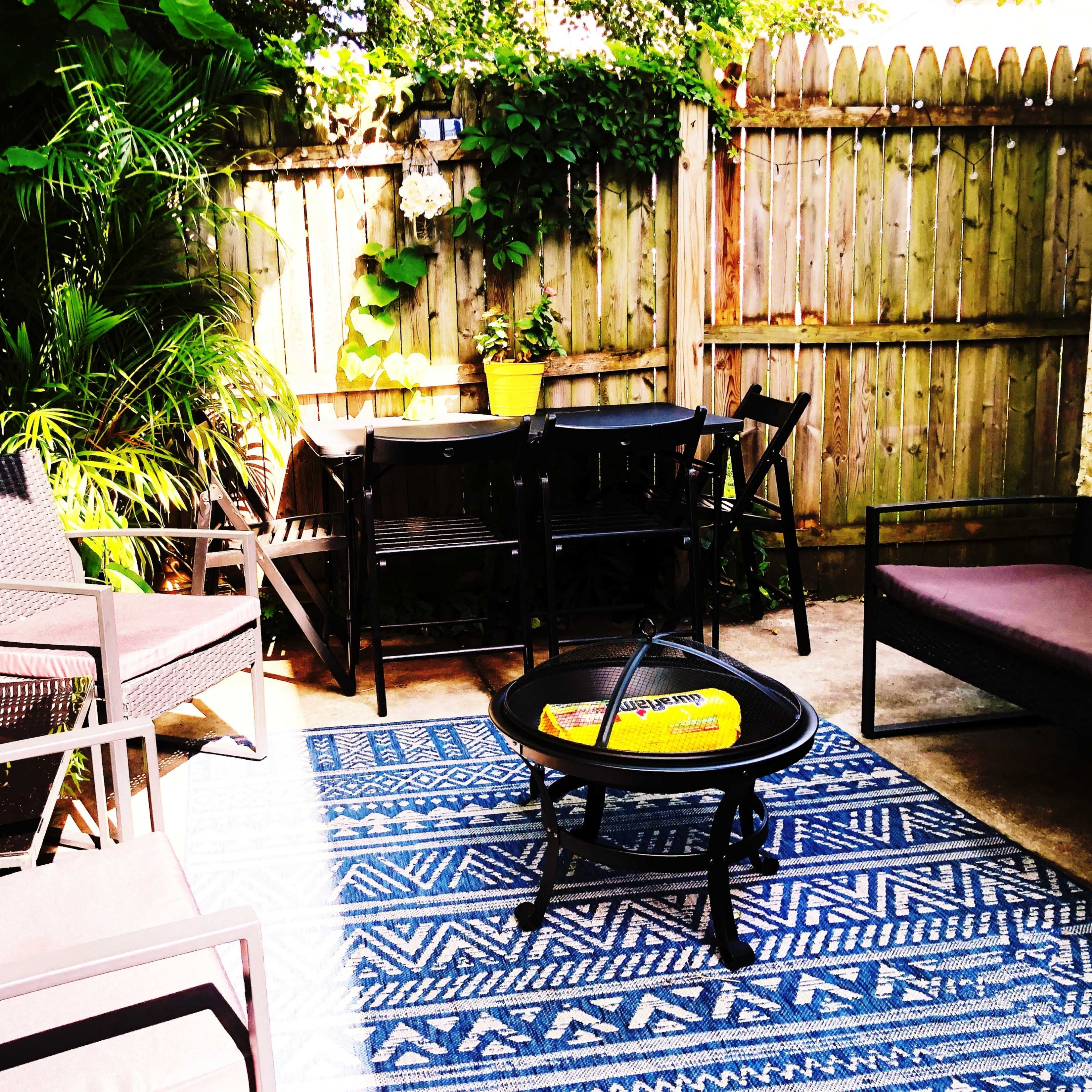 A backyard sitting area features black outdoor furniture, a patterned rug, and a fire pit surrounded by greenery and a wooden fence.