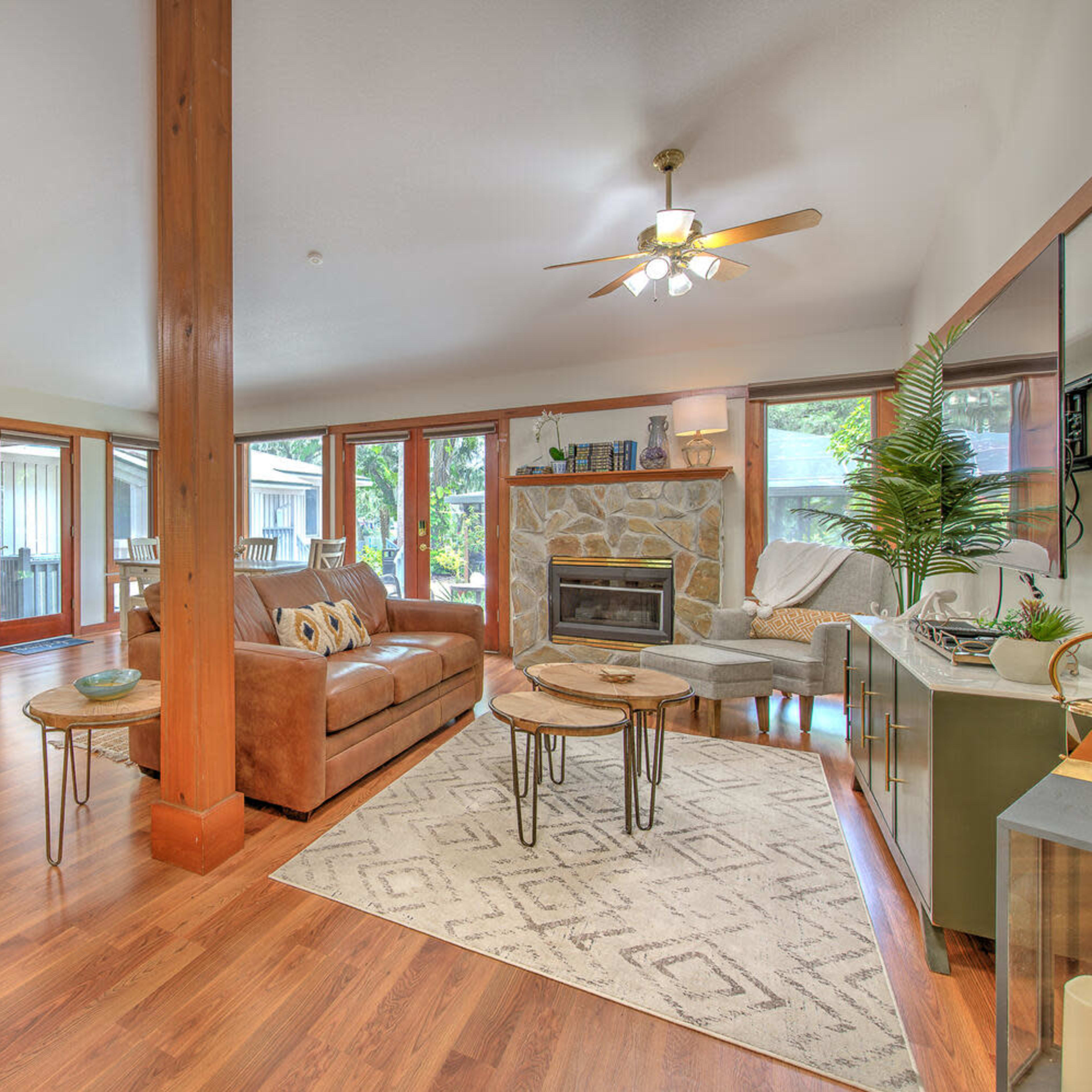 A spacious living room features a comfortable couch, a coffee table set, and a stone fireplace, illuminated by ceiling fans and large windows.