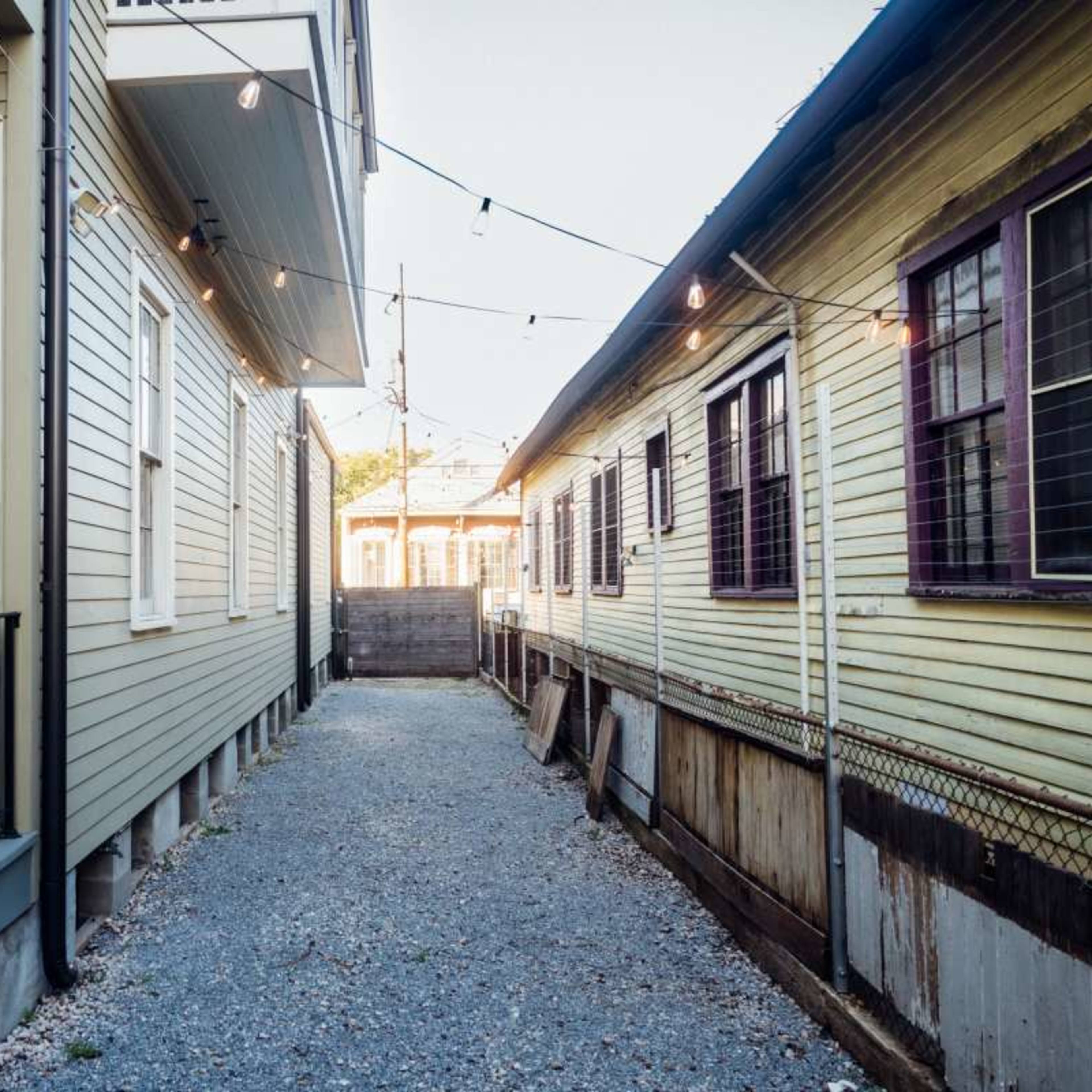 A narrow gravel pathway runs between two houses, one with a bright red door and the other with yellow siding, illuminated by string lights above.