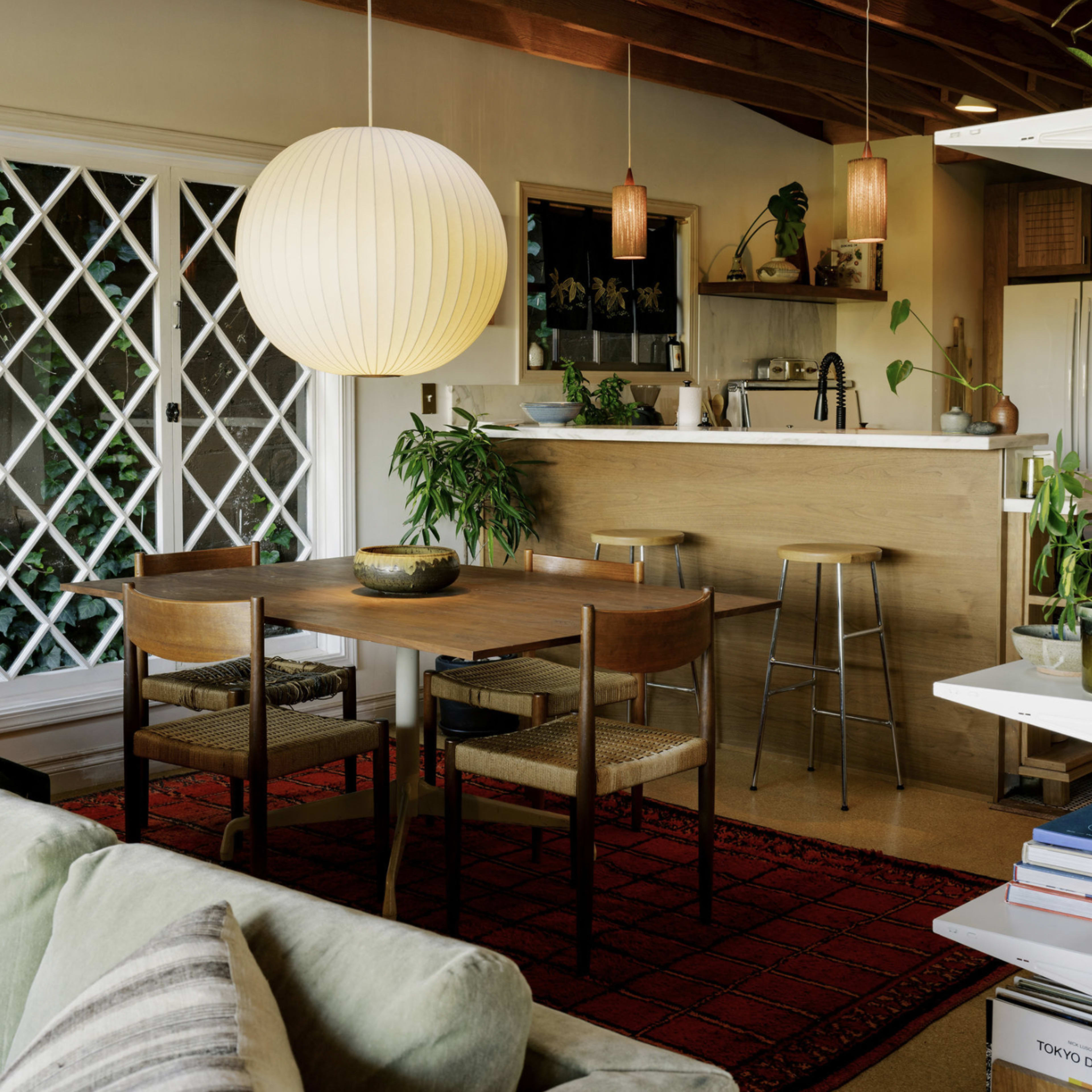 A modern, open-plan living space featuring a wooden dining table with chairs, a kitchen area in the background, and various plants throughout.