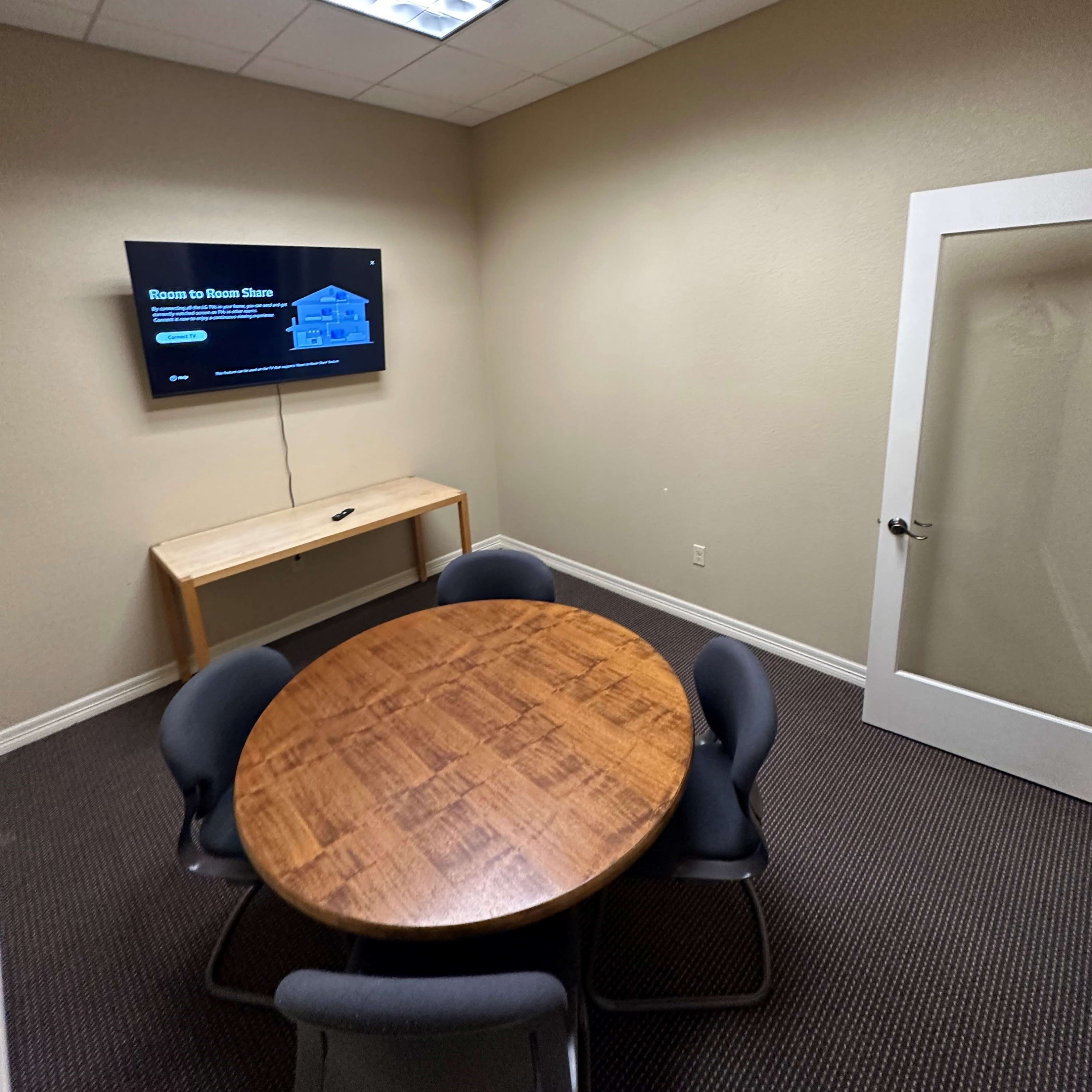 A small conference room with a round wooden table, four chairs, a wall-mounted TV, and a glass door.