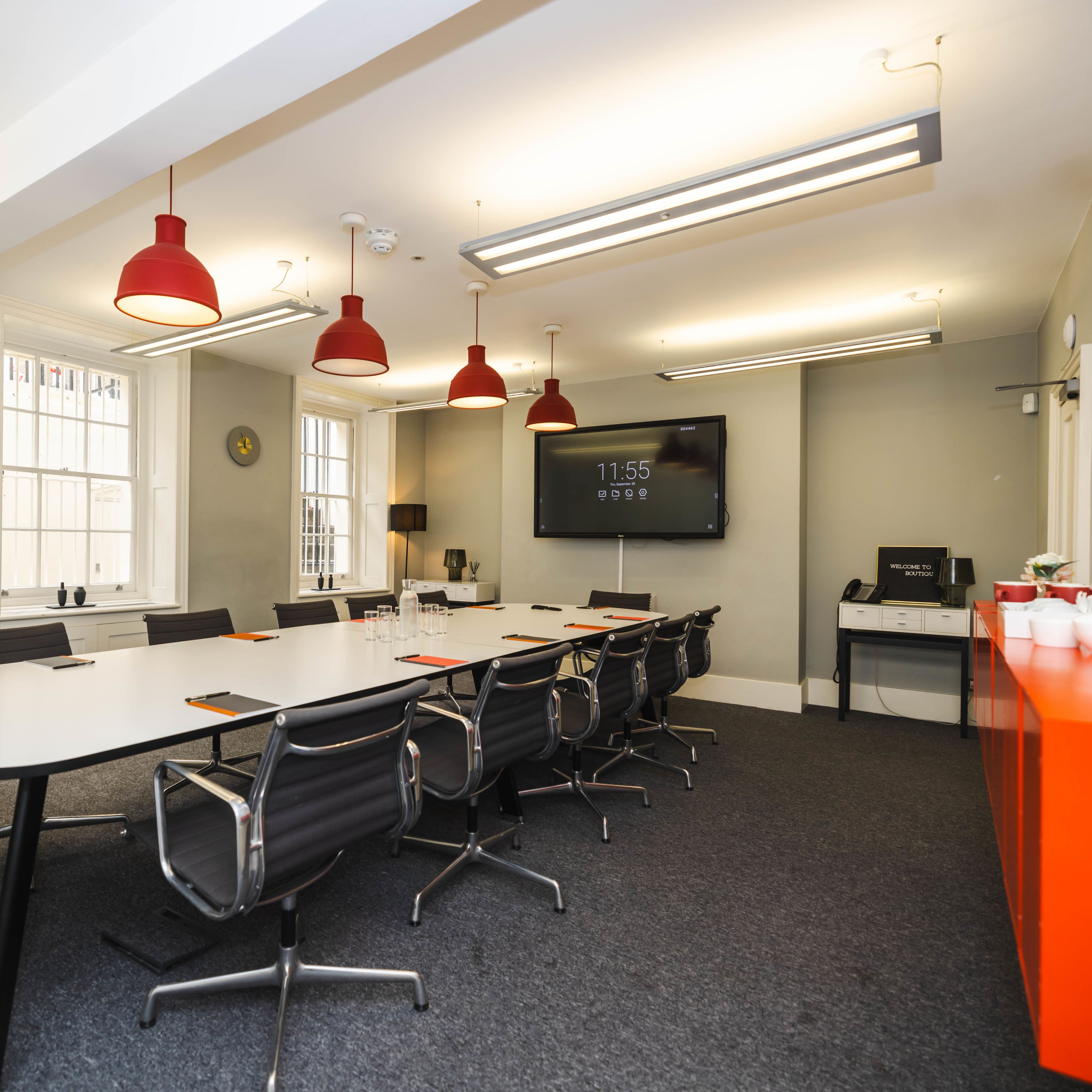 A modern conference room with a long white table, black chairs, red pendant lights, and a large screen displaying the time on one wall.