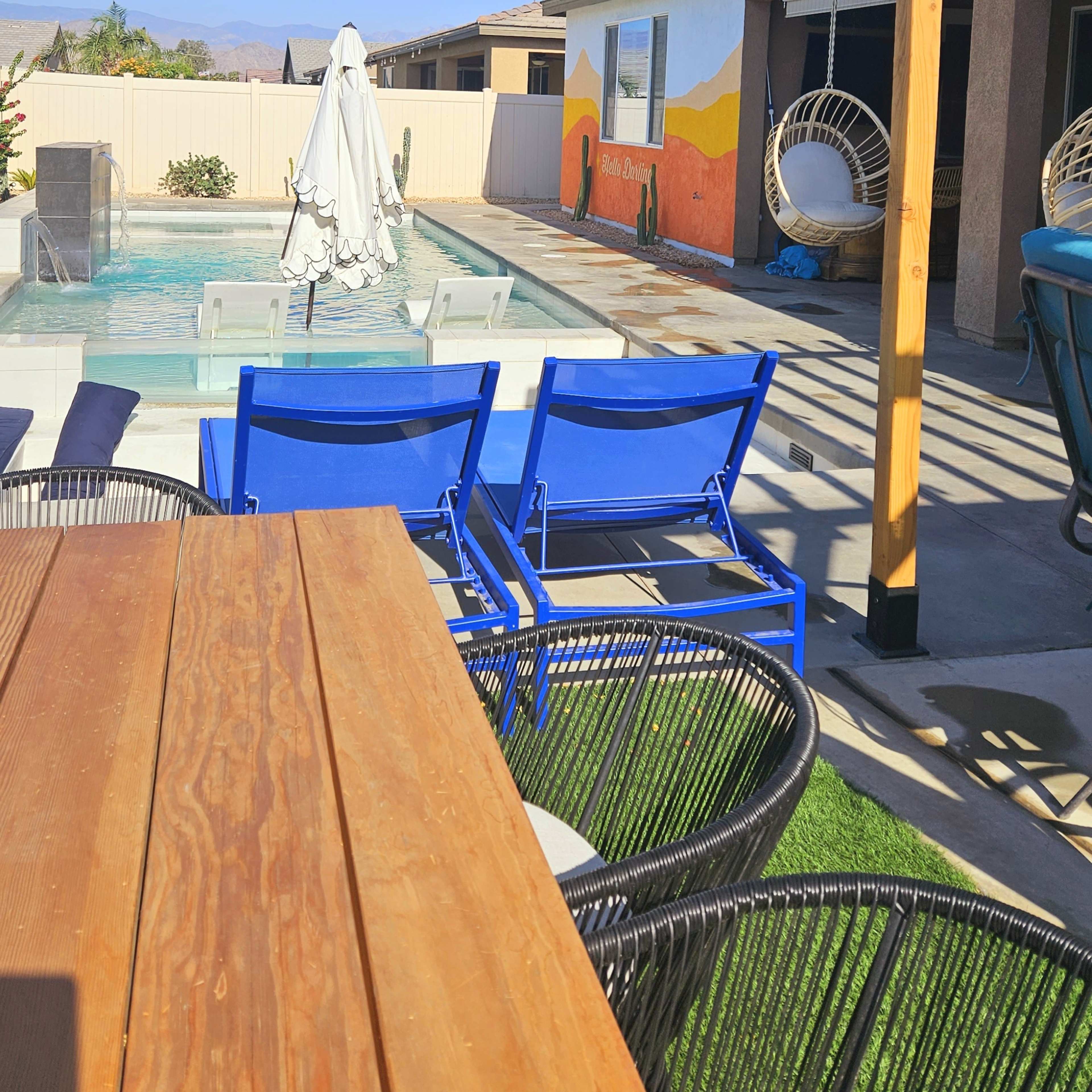 A backyard pool area with blue chairs, a wooden table, and a house featuring a mural in the background.