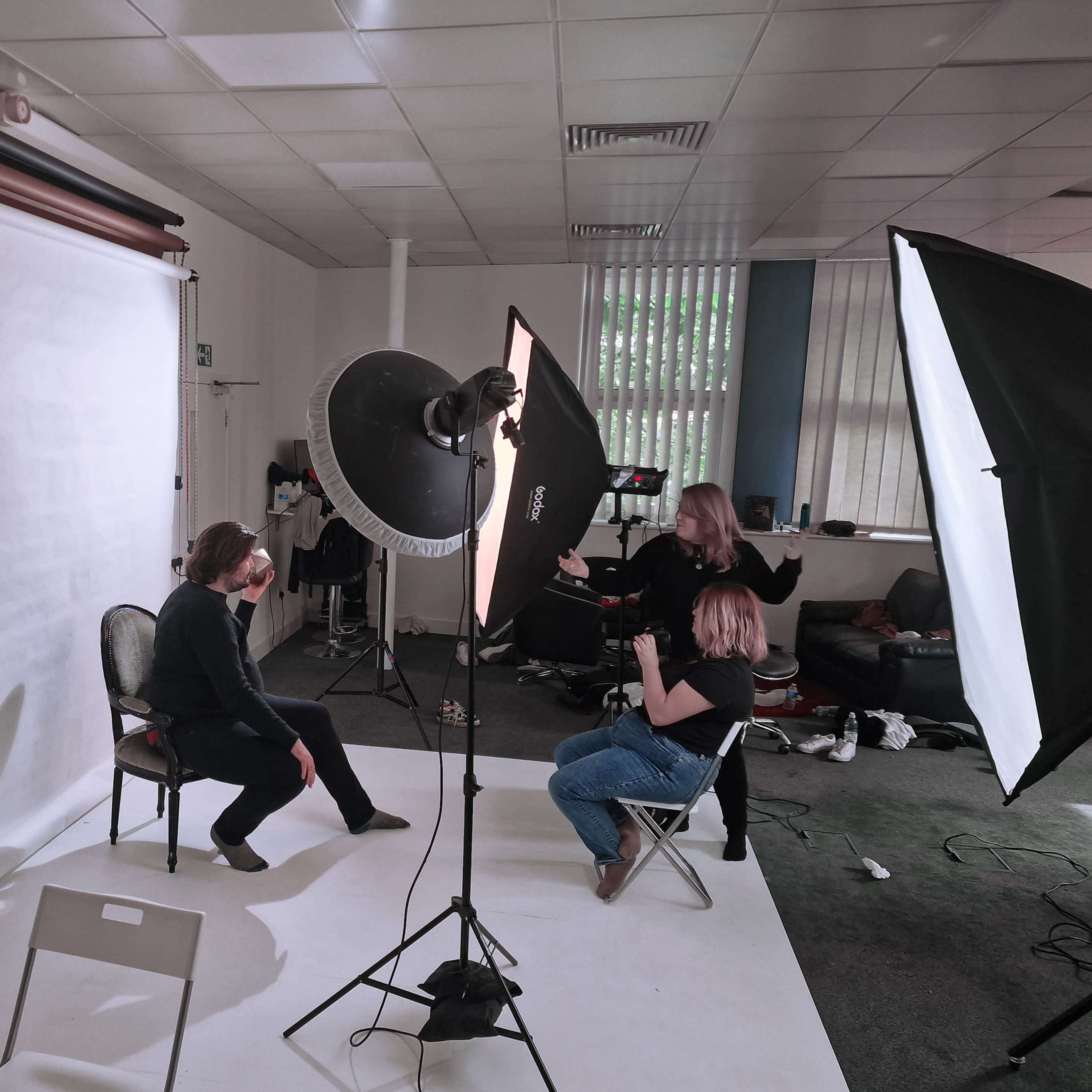 A person sits on a chair in front of a backdrop while another person photographs them, surrounded by lighting equipment in a studio.