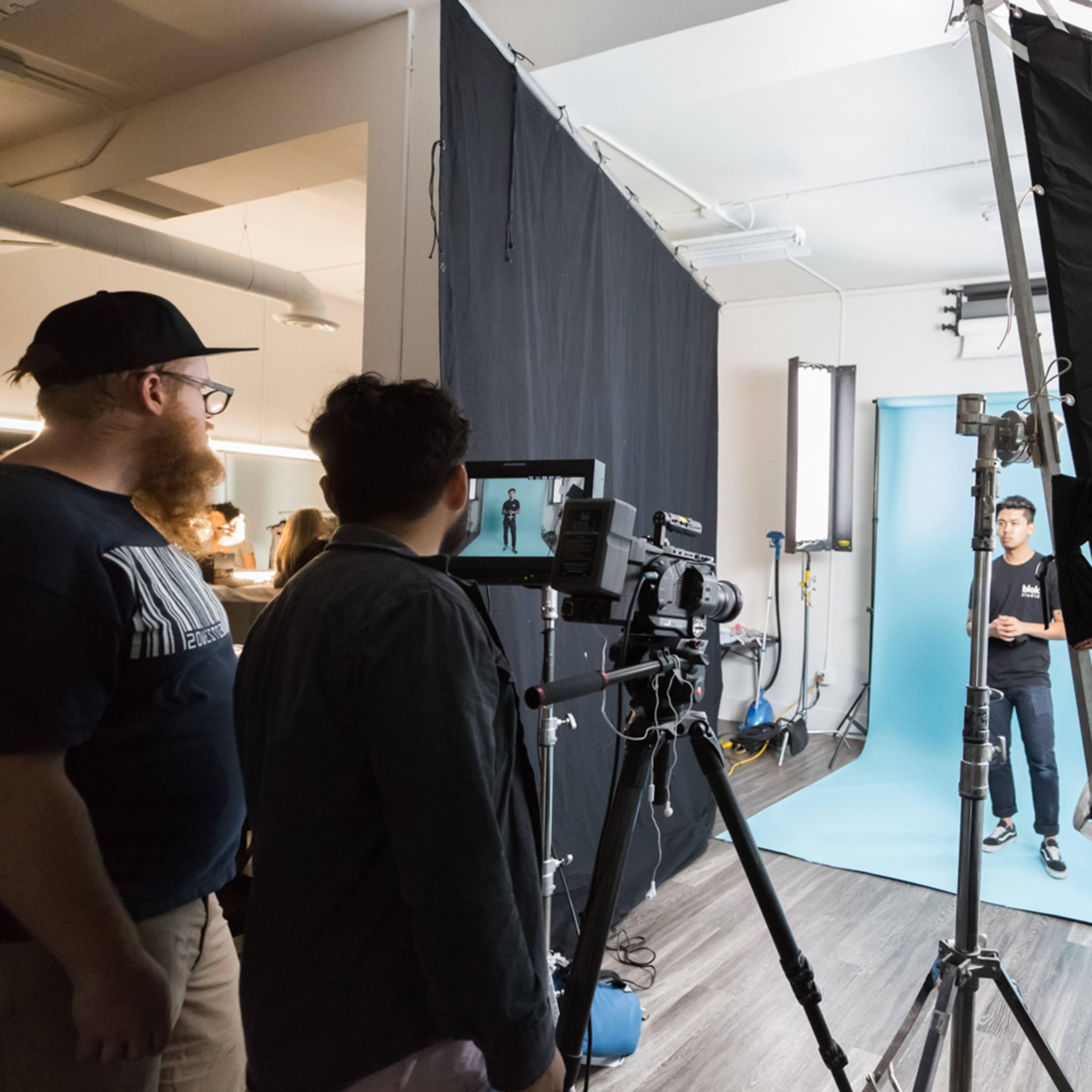A film crew operates equipment while a person stands against a light blue backdrop in a studio setting.
