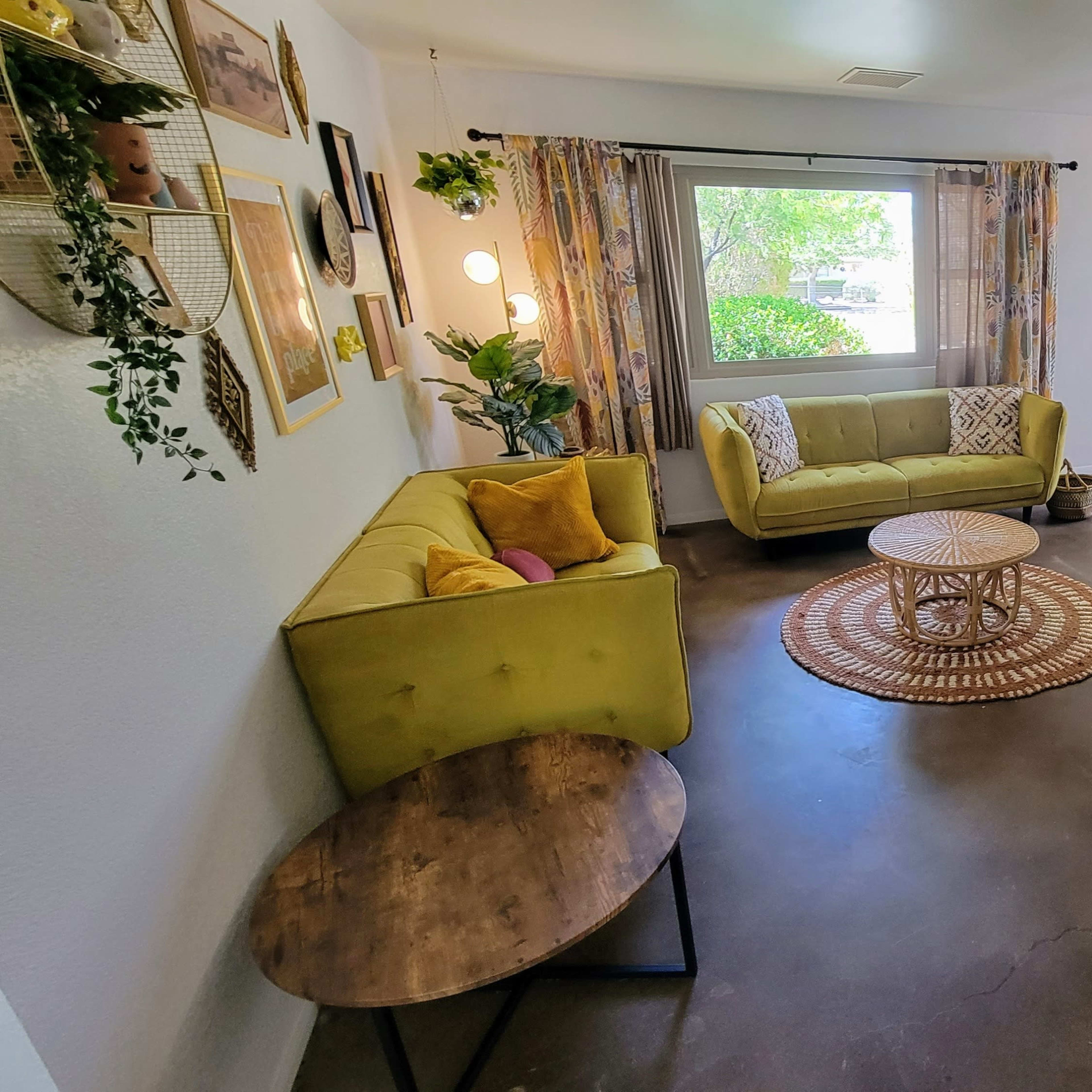 A bright living room features two yellow sofas, a round rug, and a wooden coffee table beside a window with patterned curtains.