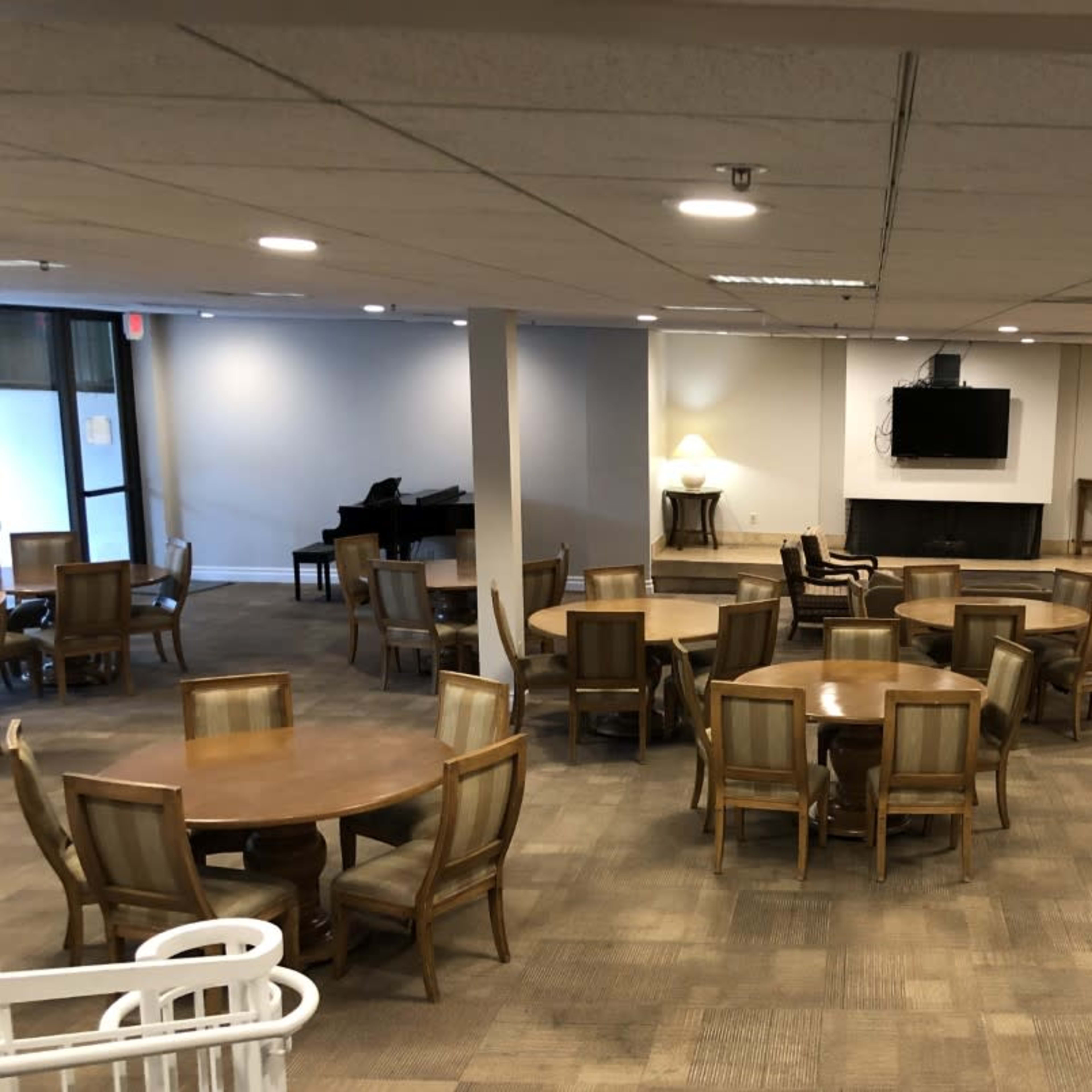 A large common area with several round tables and chairs, a piano in one corner, a television on the wall, and a kitchenette area.
