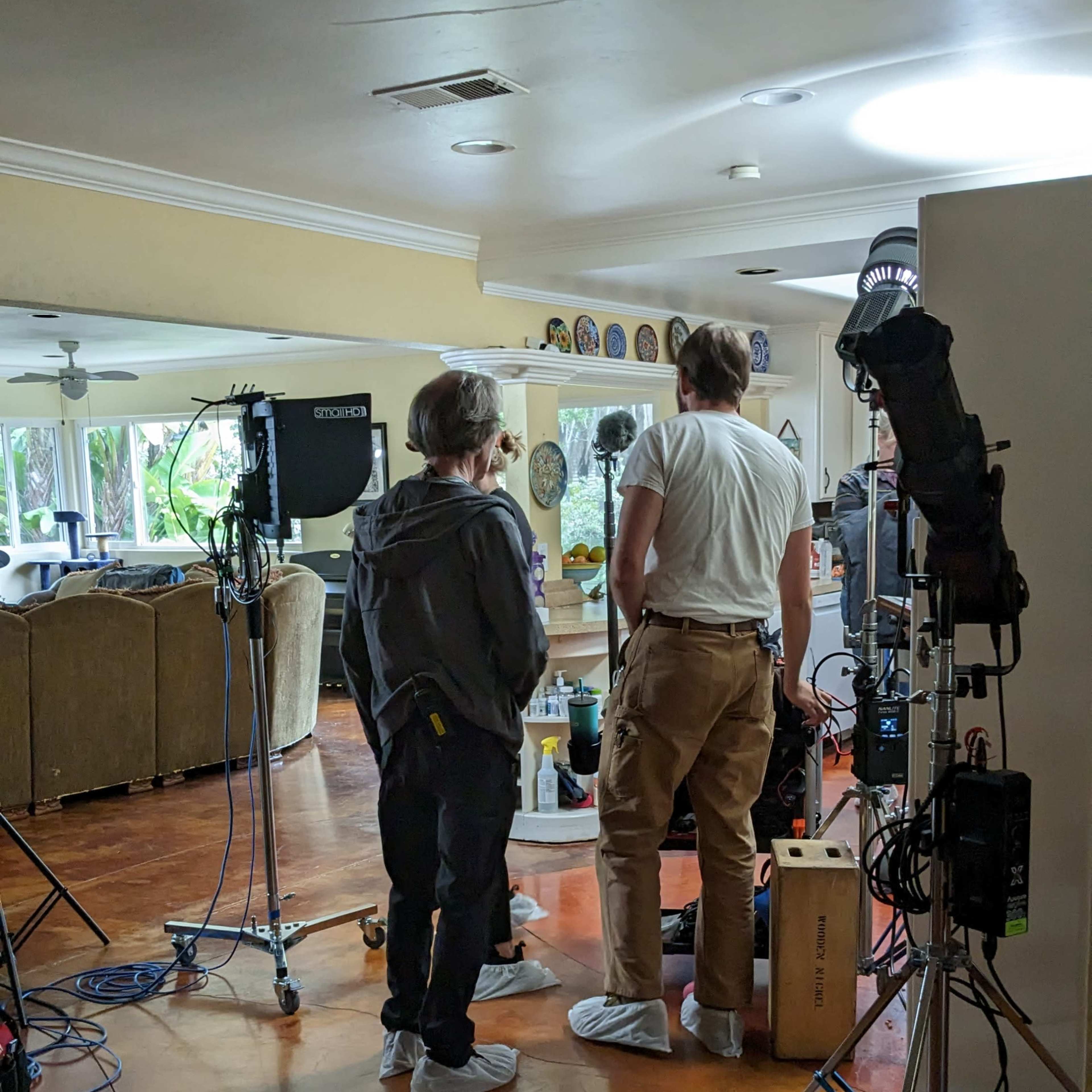 Two crew members prepare equipment in a living room set for a film shoot, while cameras and lighting gear are arranged nearby.