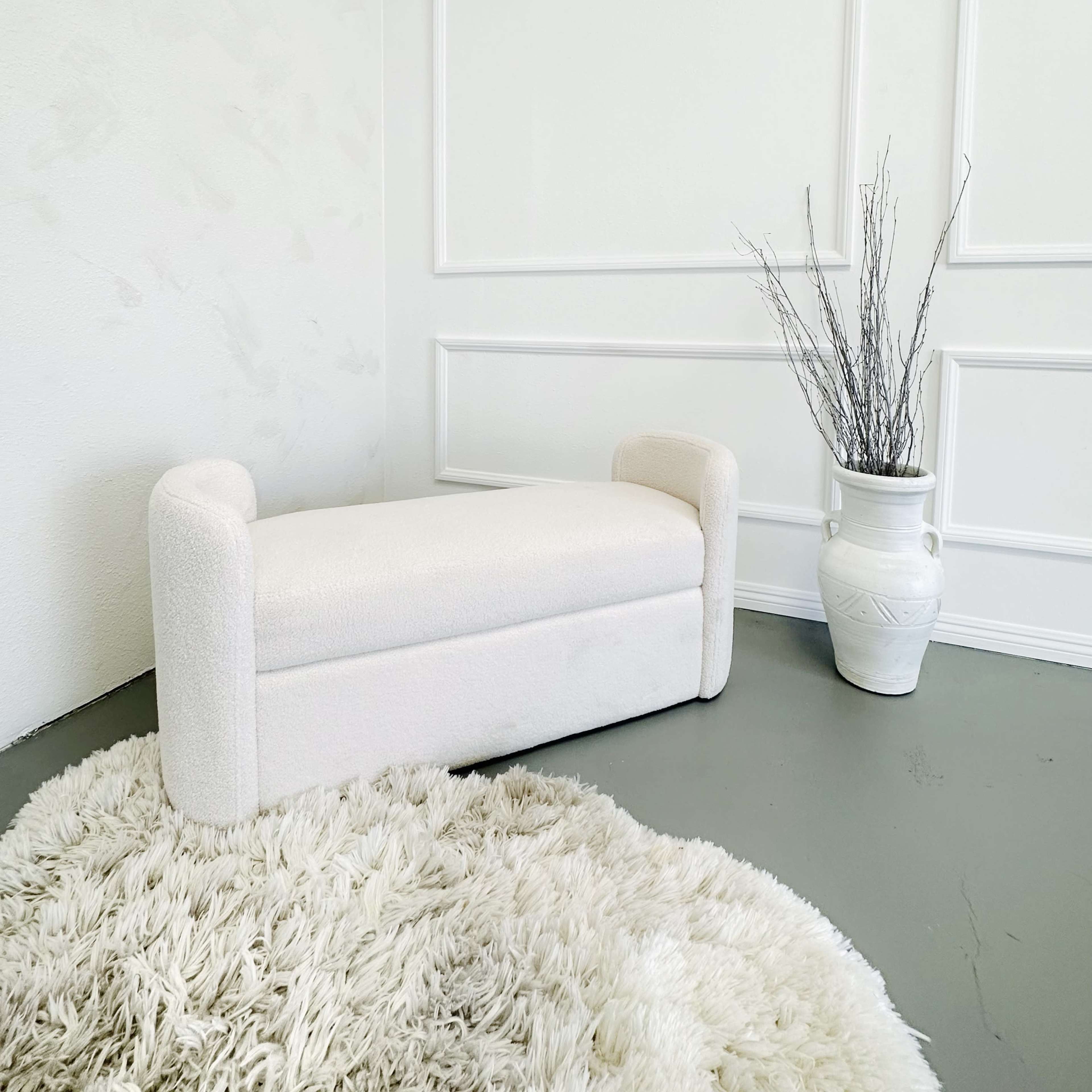 A white upholstered bench sits next to a decorative vase with dried branches, on a soft, round rug in a minimalist room.