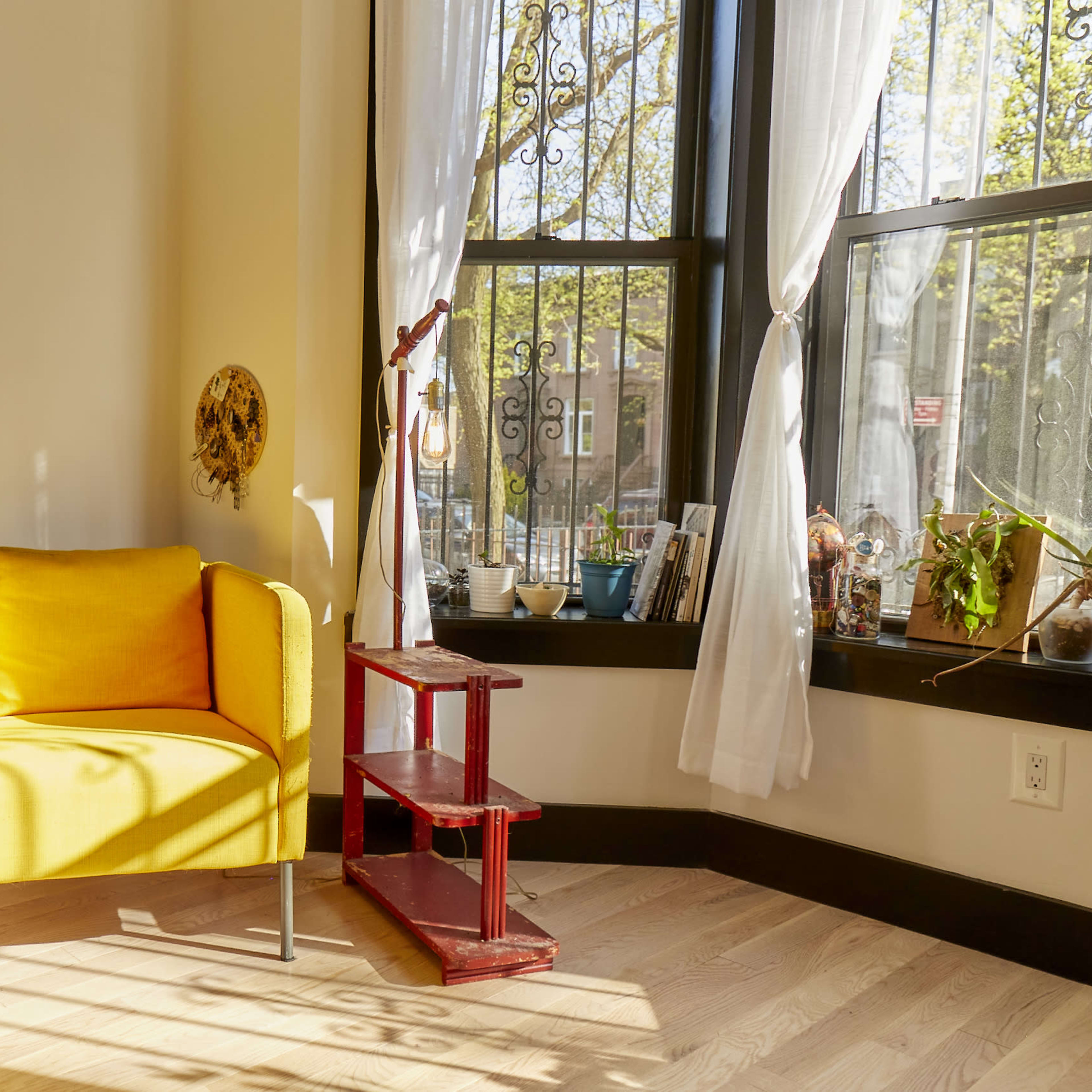 A yellow armchair is positioned beside a window with flowing white curtains, next to a potted plant and a red wooden shelf displaying various items.