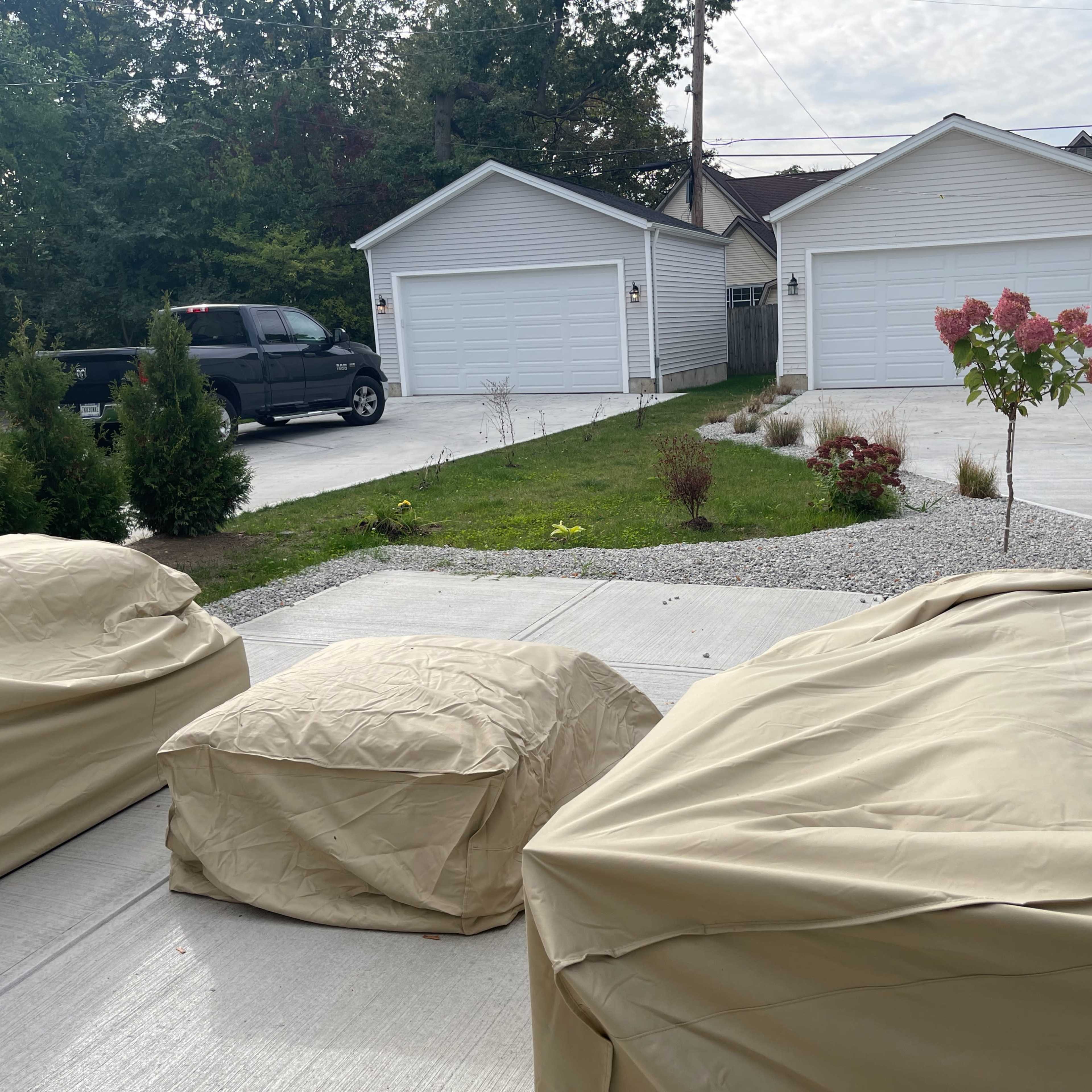 A driveway with three covered items in the foreground, leading to two garages in the background.