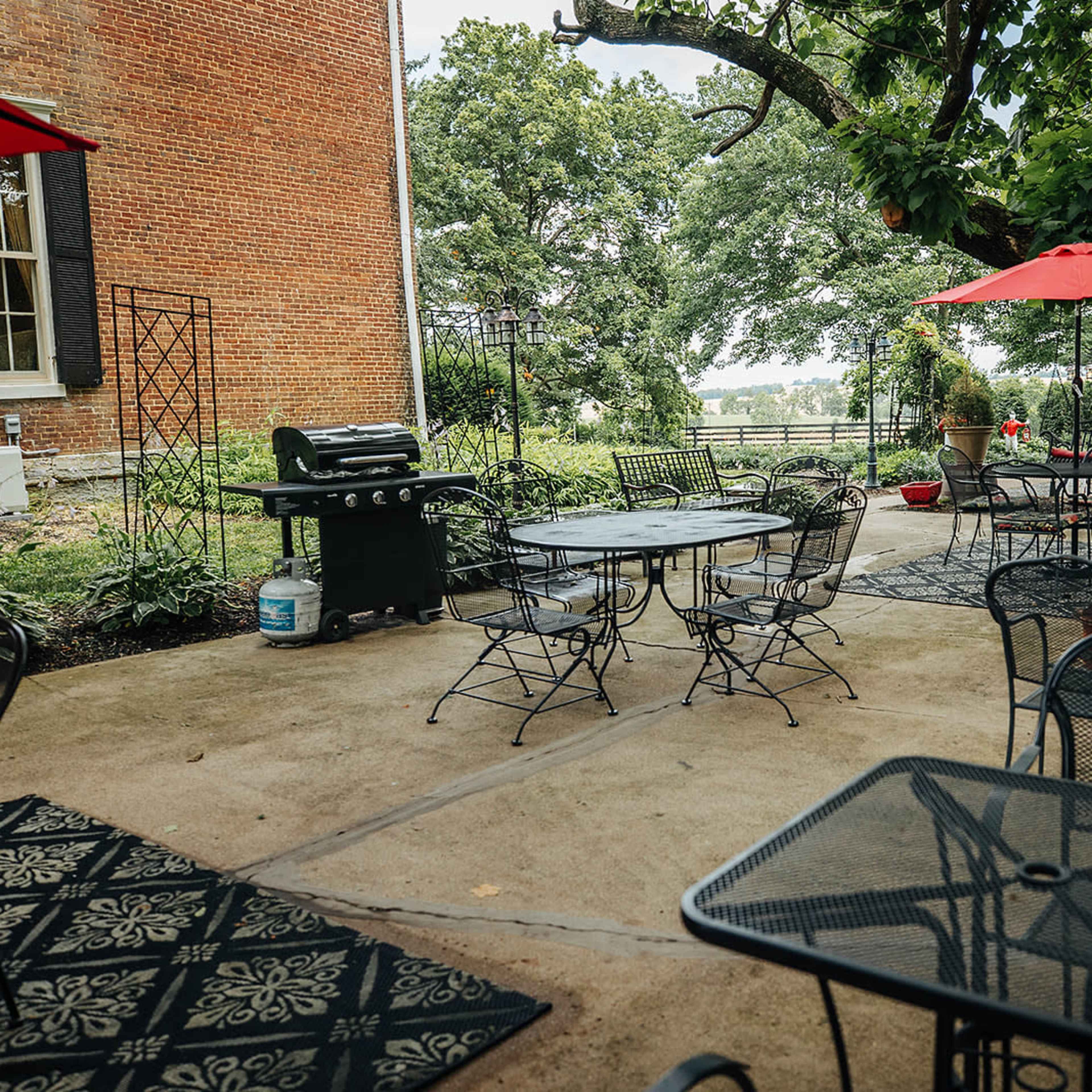 A patio area features metal tables and chairs surrounded by greenery and umbrellas, adjacent to a brick building.