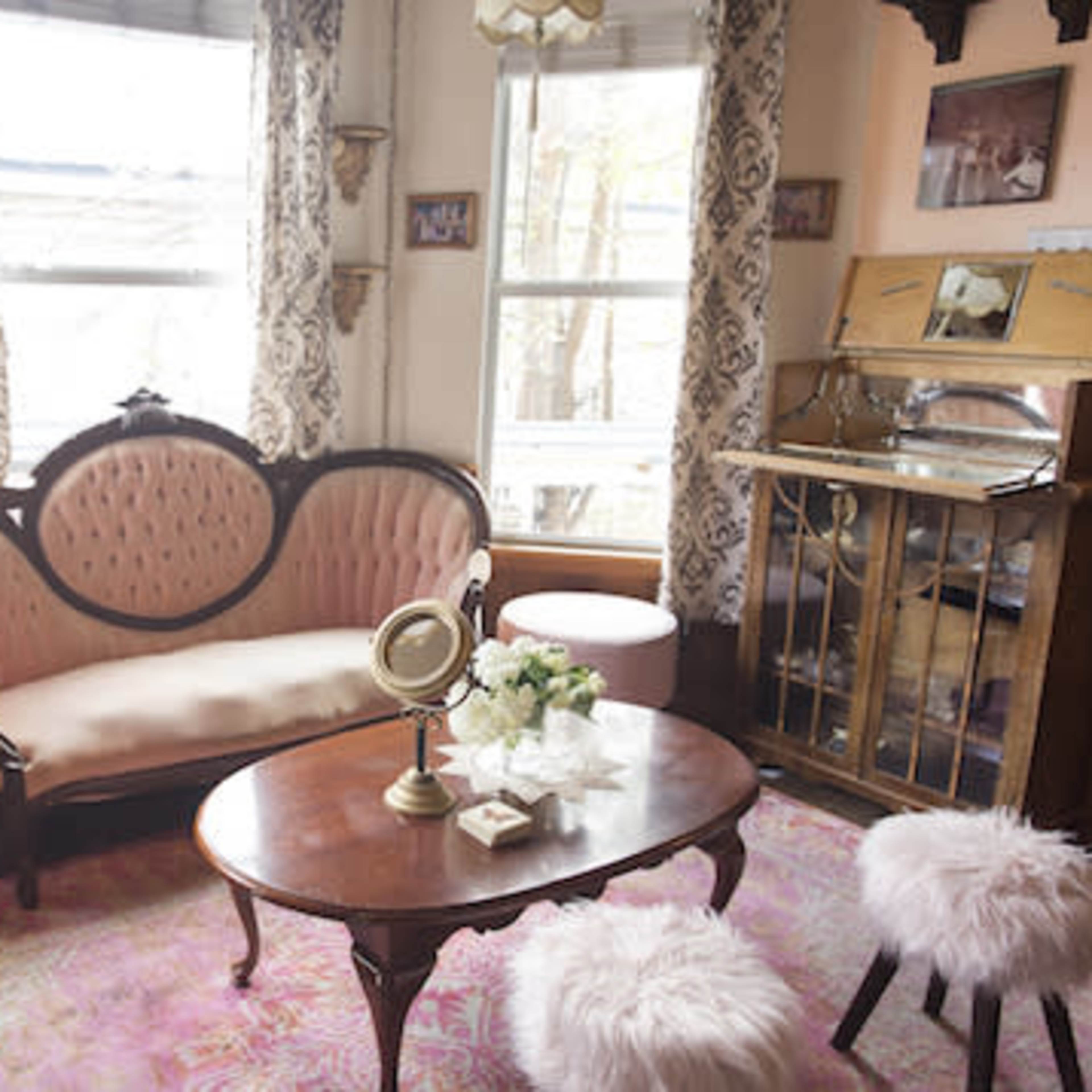 A vintage sitting room features a plush pink sofa, a wooden coffee table, and a decorative cabinet, illuminated by natural light from the windows.