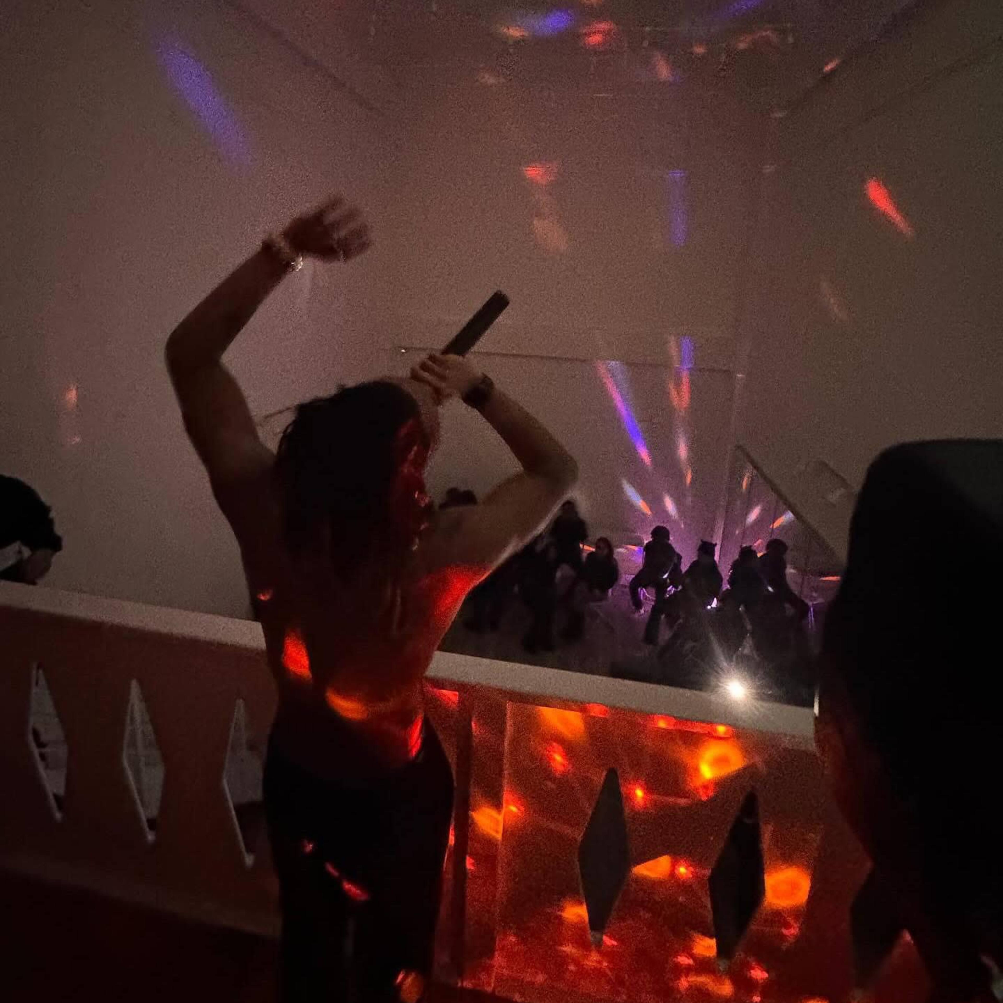 A person with an outstretched arm holds a microphone on a staircase as colorful lights illuminate a crowded room below.