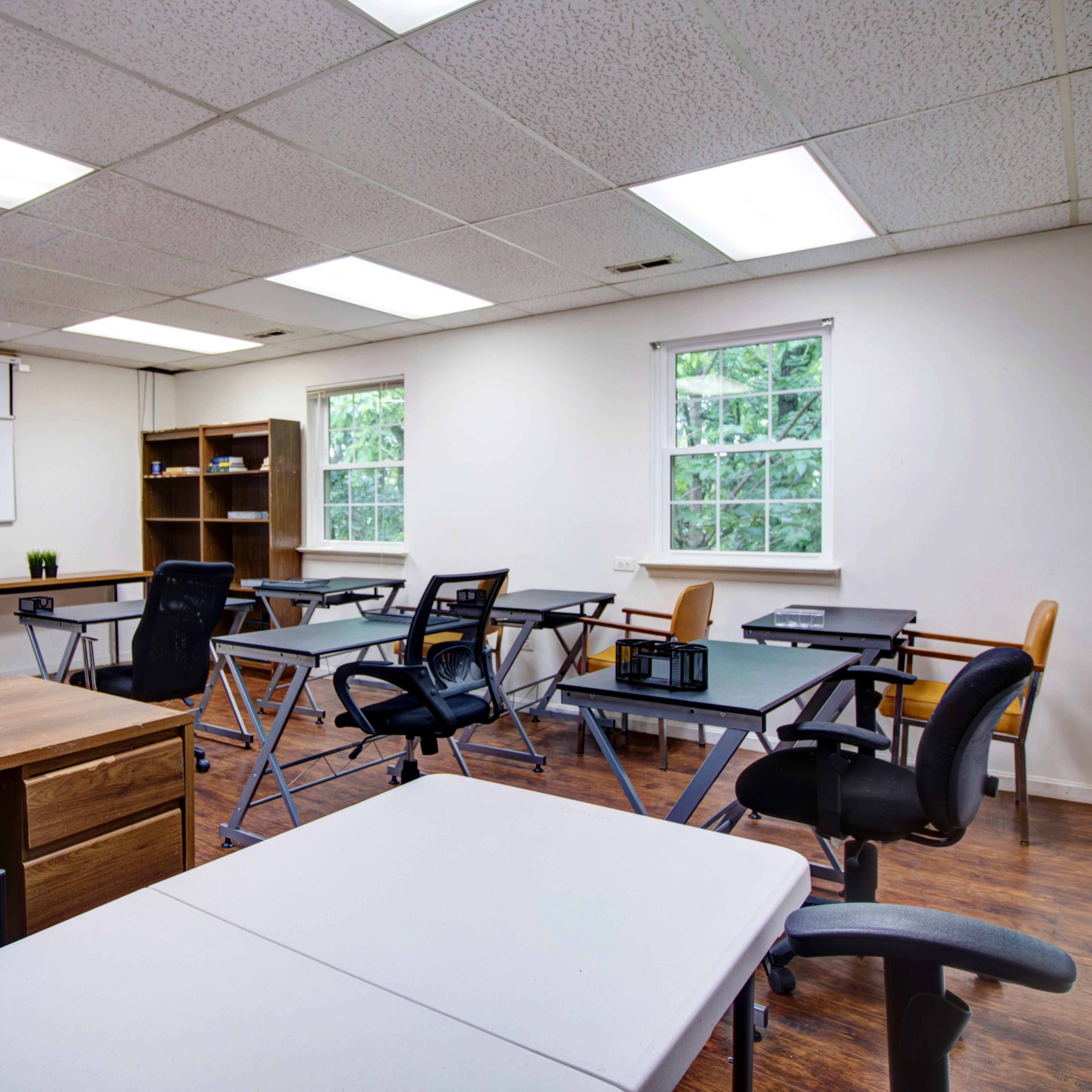 A well-lit classroom with multiple desks, chairs, a whiteboard, and shelves against the walls.