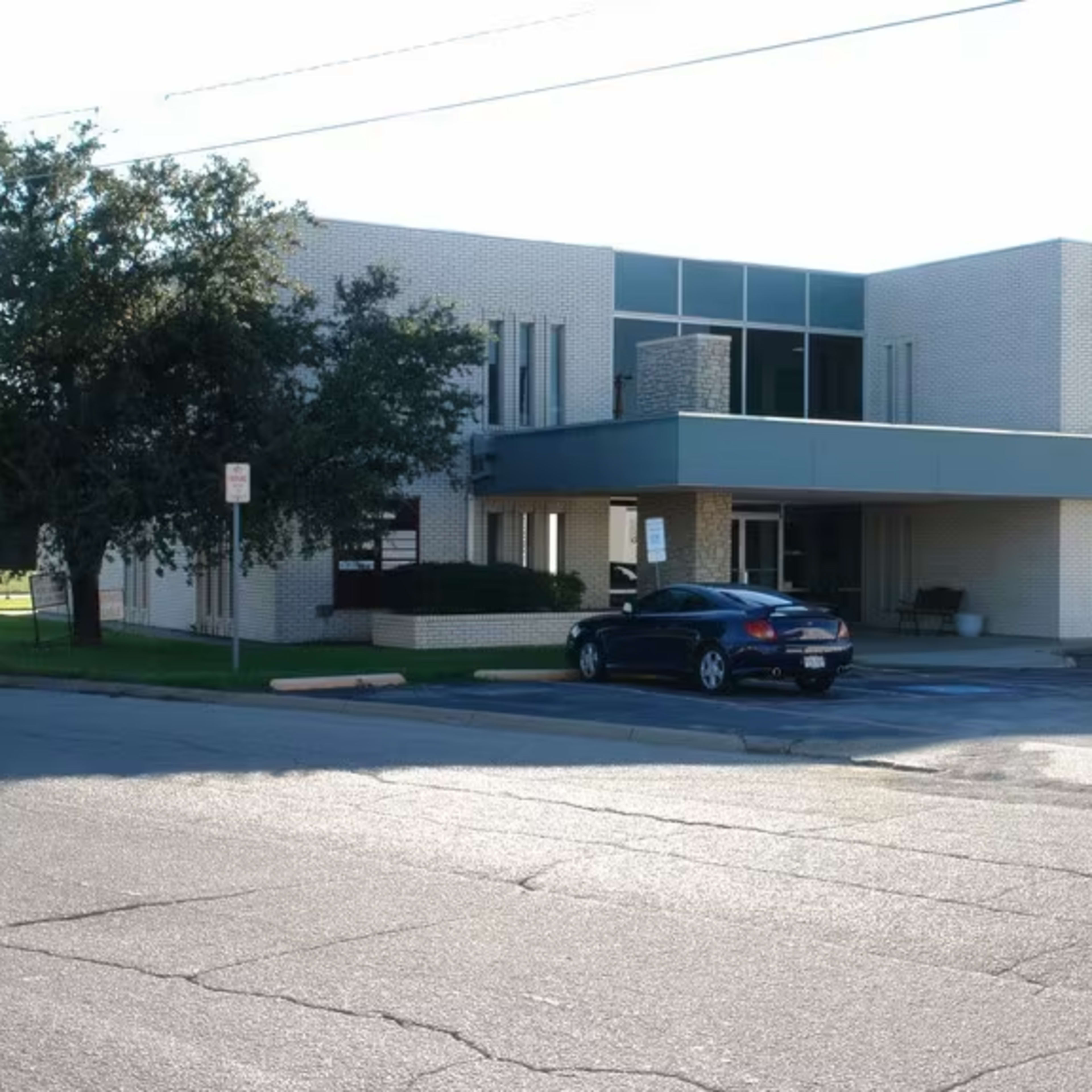 A two-story commercial building with a light-colored brick facade, featuring several windows and two parked cars in the foreground.