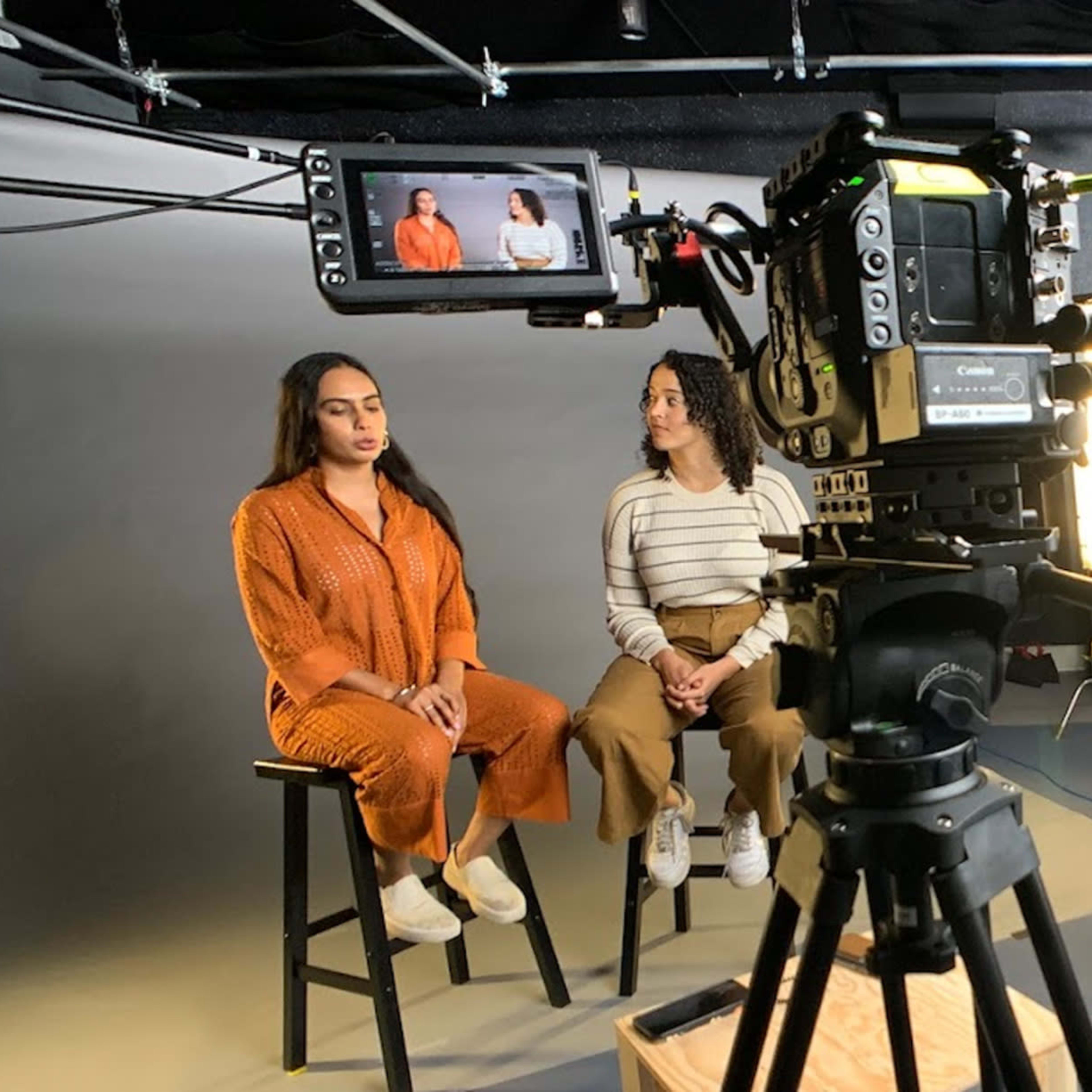Two women are seated on stools in a studio setting while a man operates a camera on a tripod.