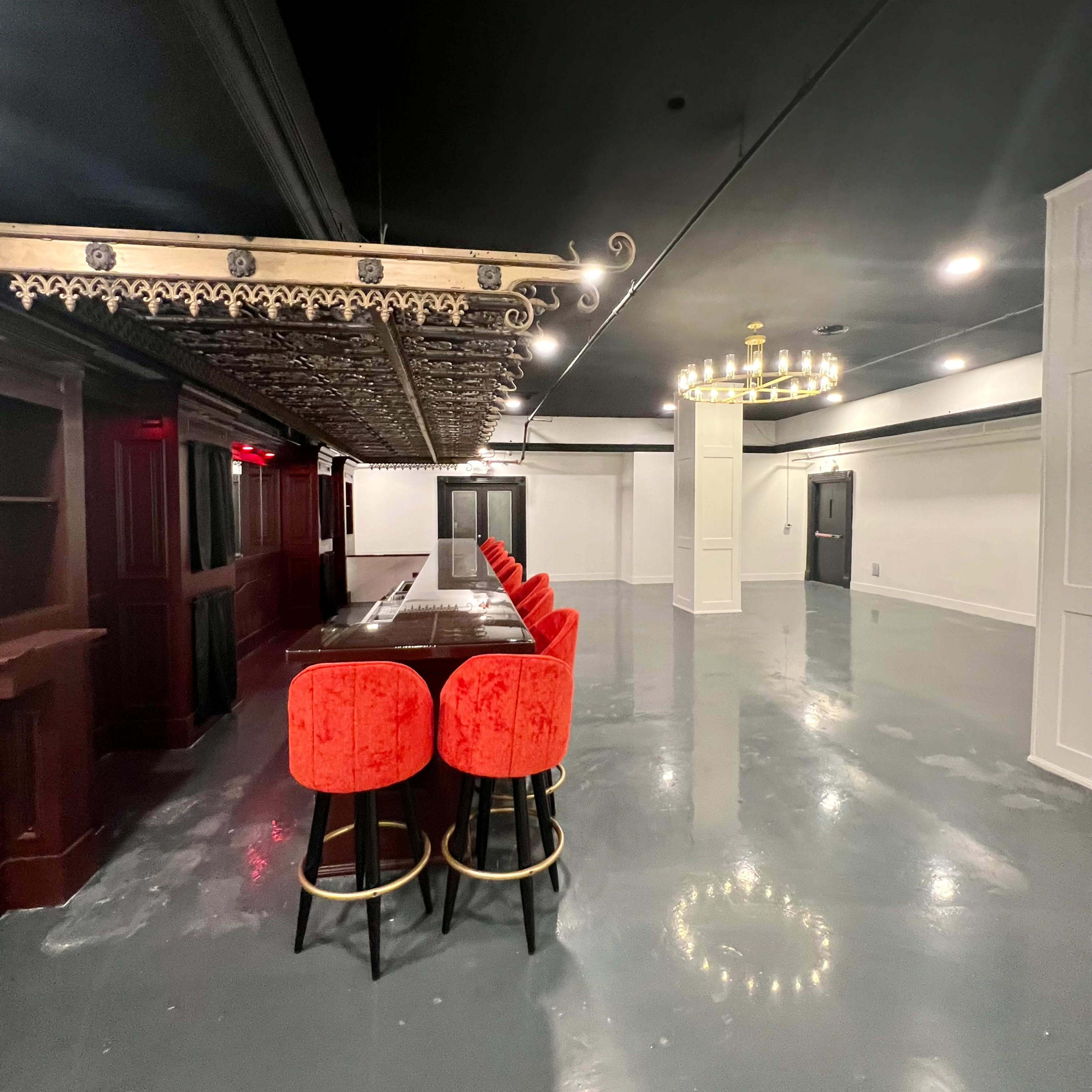 A spacious room with a long dark table, red upholstered stools, ornate ceiling details, and a chandelier.