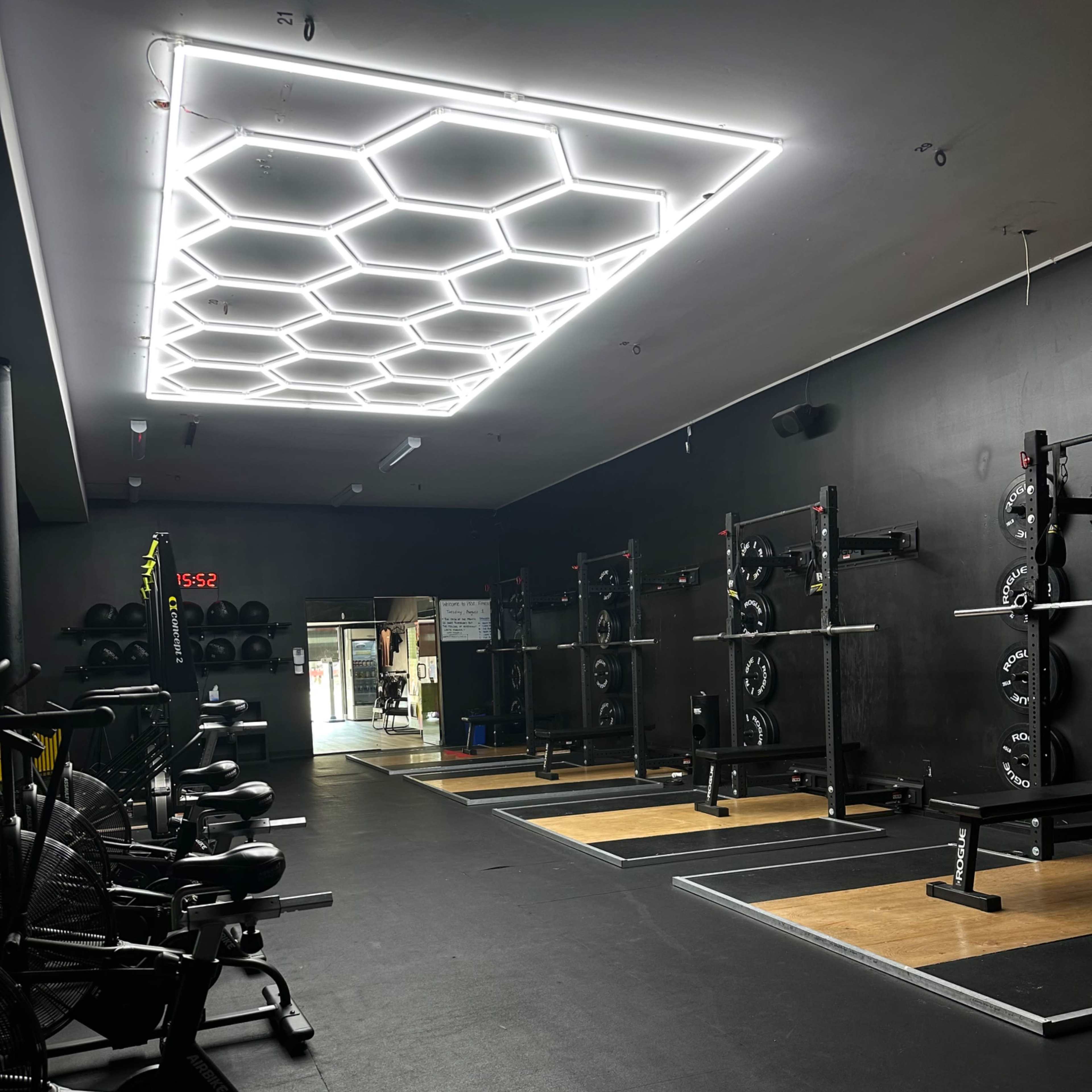 A gym interior with workout stations, including weightlifting racks and exercise bikes, illuminated by a hexagonal LED ceiling light.