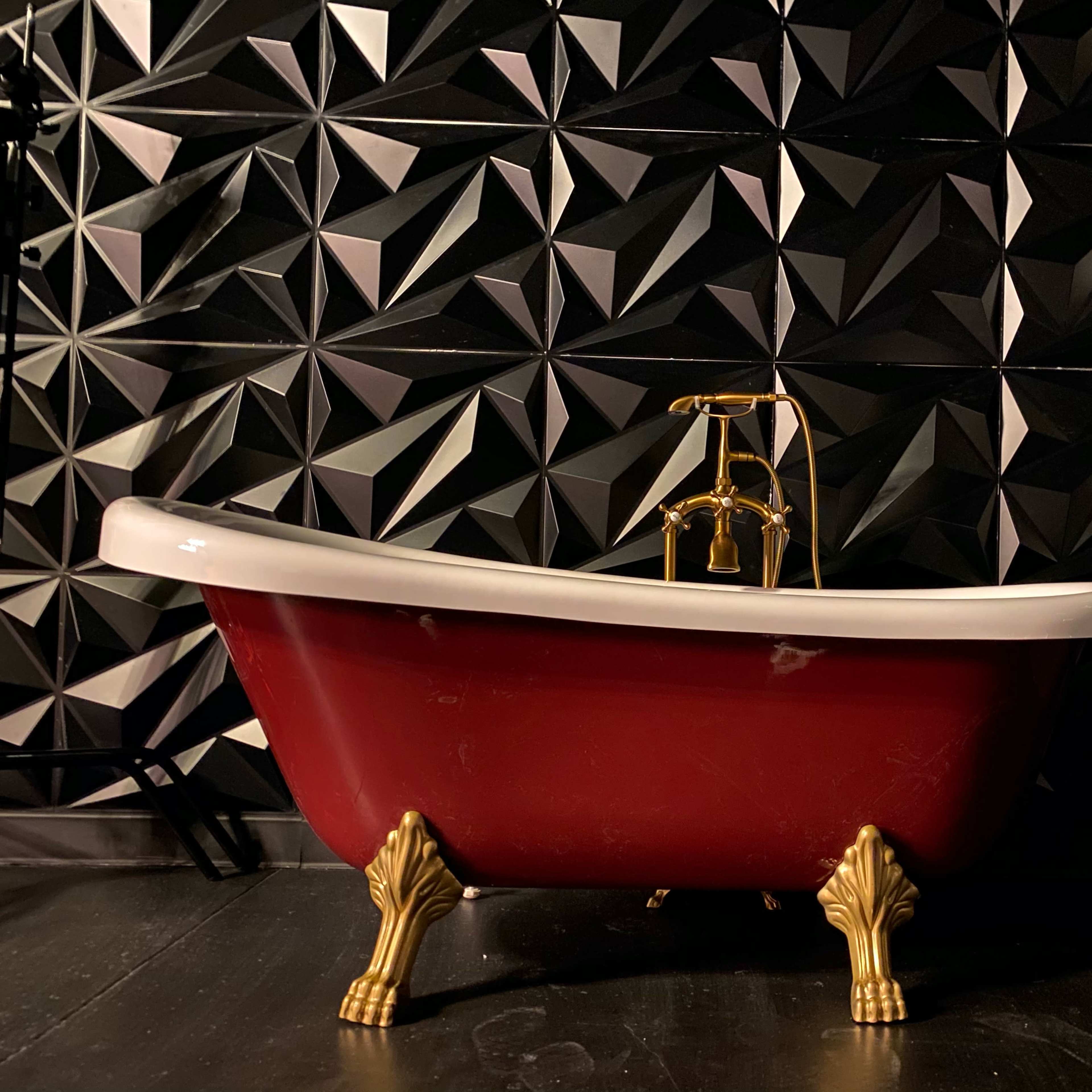 A vintage red bathtub with gold fixtures sits against a black geometric textured wall.