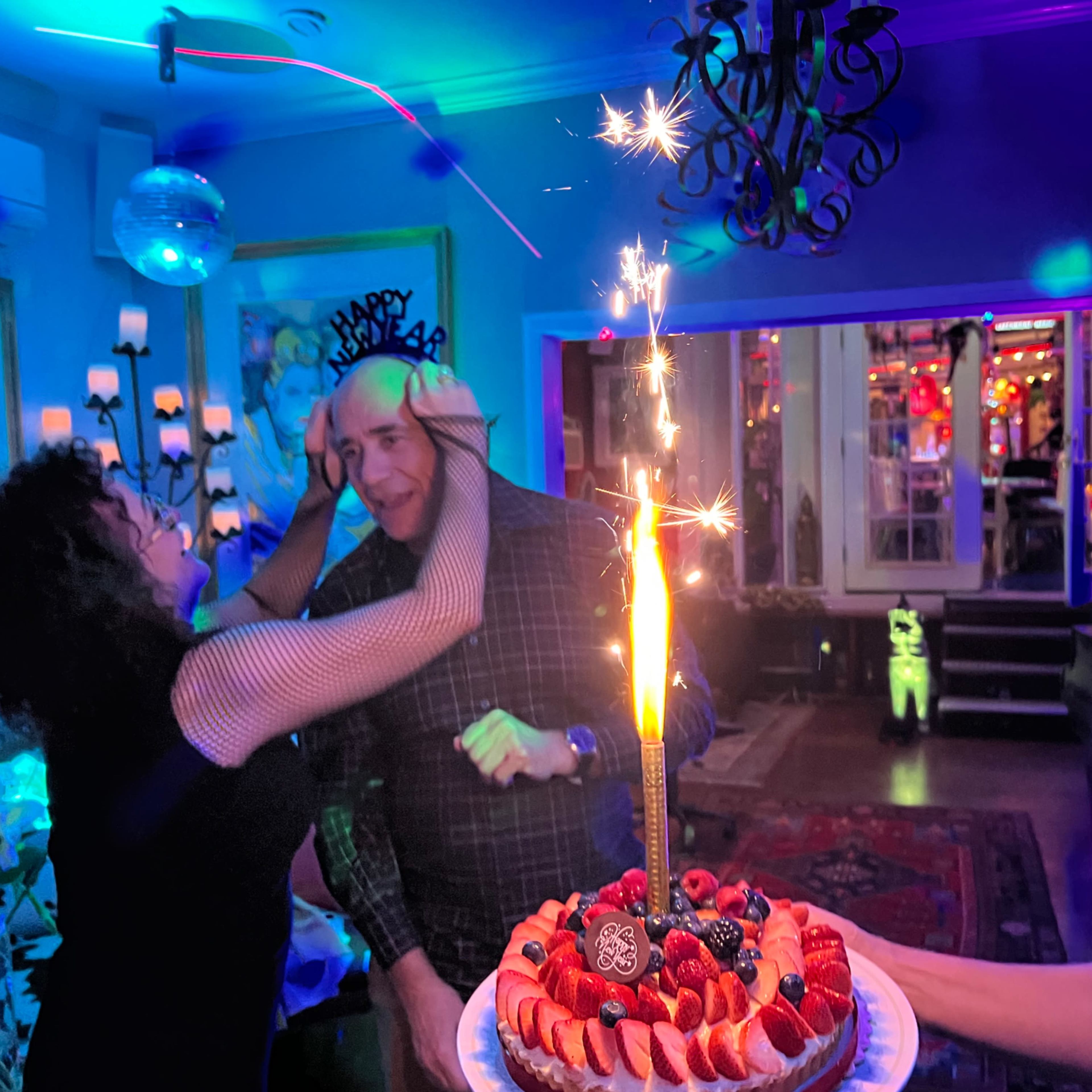 A person celebrates a birthday with a decorated cake topped with a lit sparkler while guests enjoy the party atmosphere filled with colorful lights.
