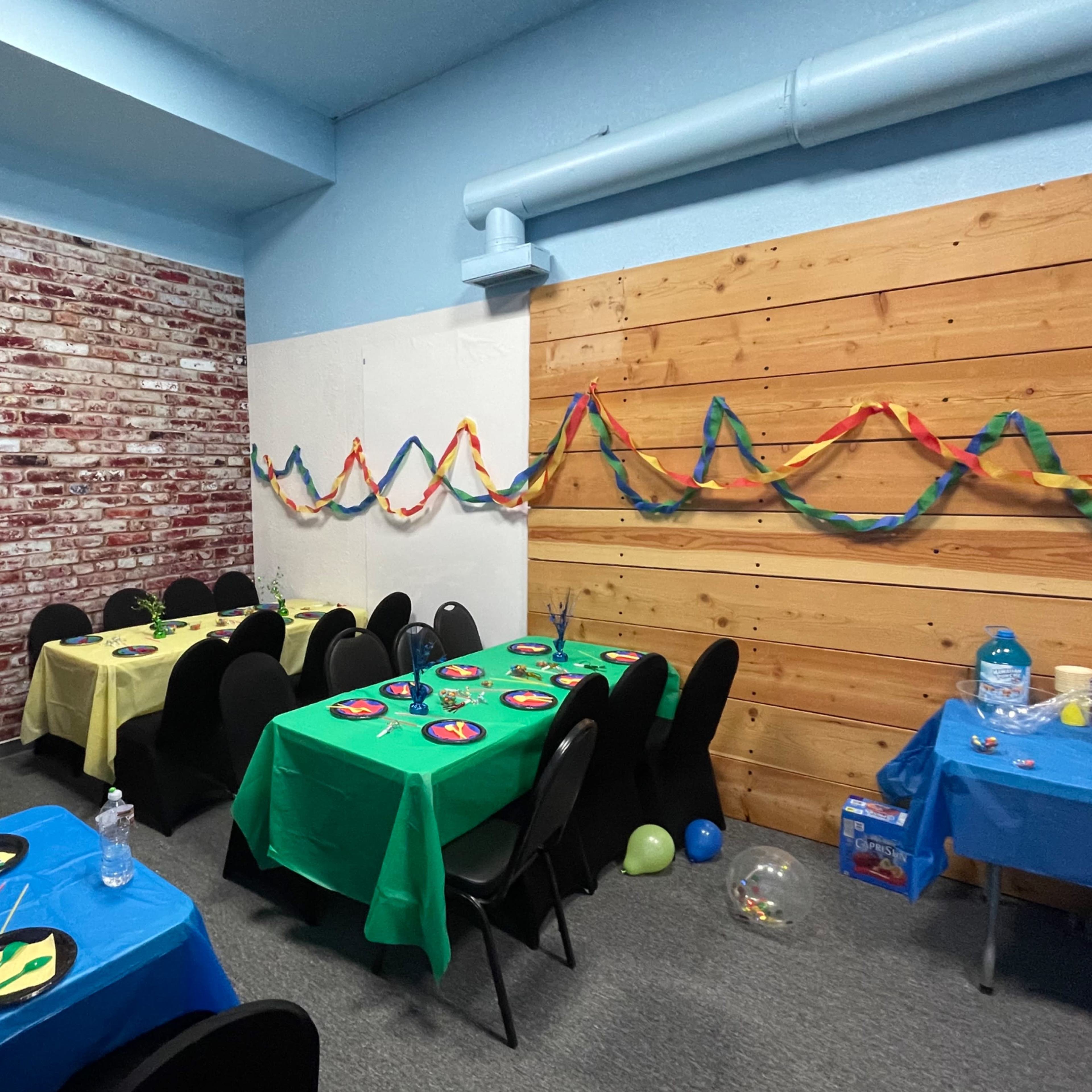 A decorated room with tables covered in bright tablecloths, featuring colorful decorations and party supplies against a backdrop of a brick wall and wooden paneling.