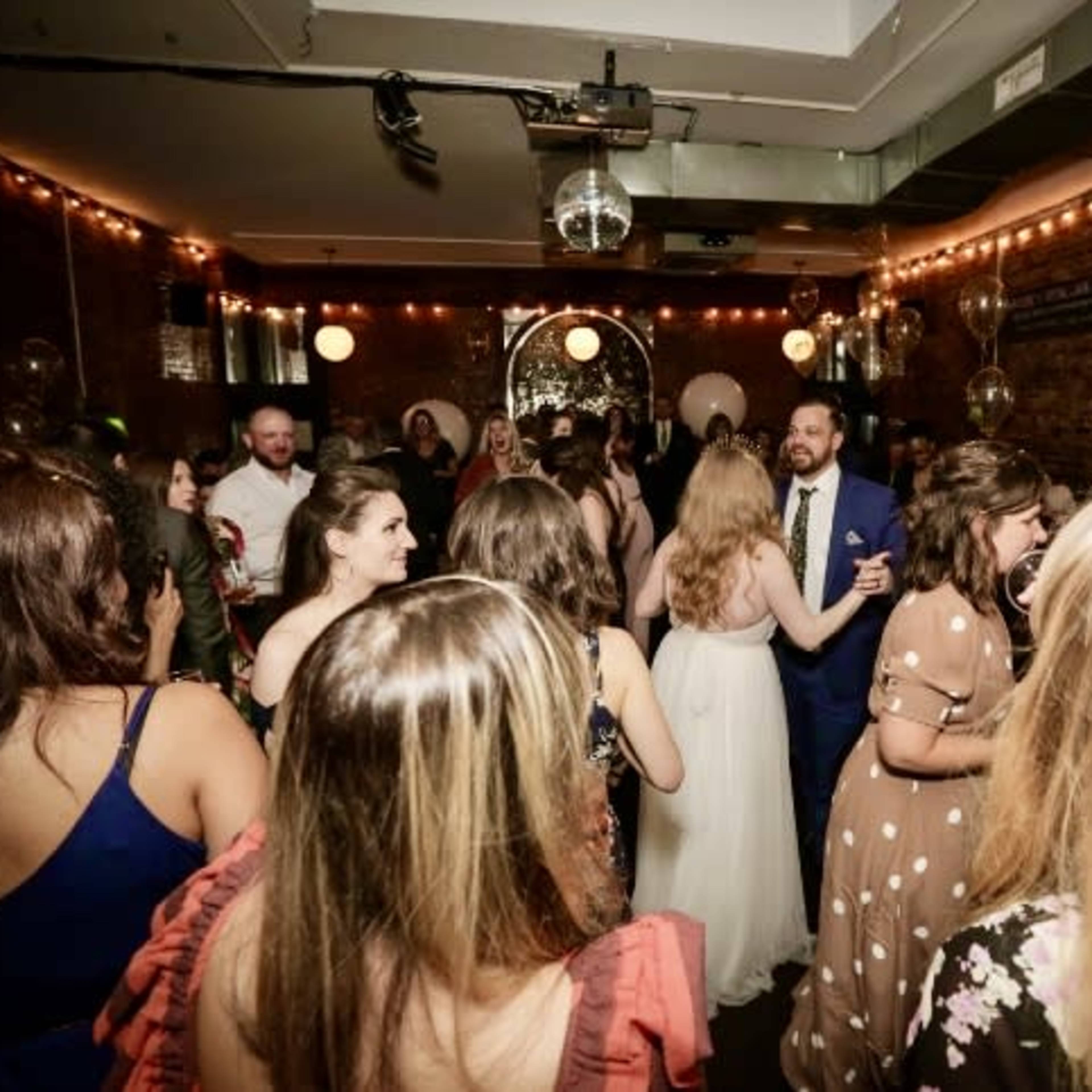 A crowd of people dances at a lively indoor celebration adorned with string lights and decorative balloons.