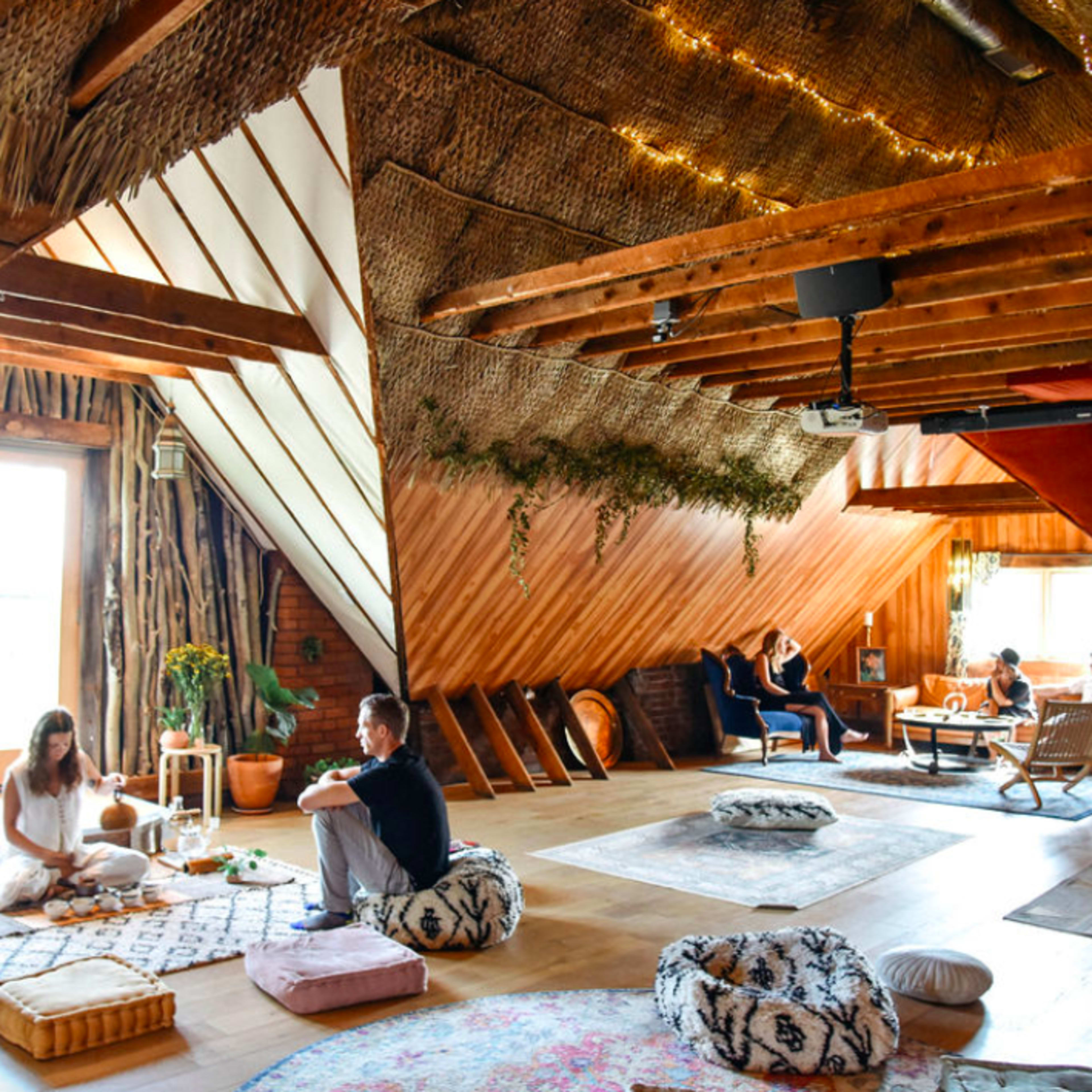 The scene features a spacious, wooden loft filled with people engaged in various activities, including lounging, socializing, and having tea, with decorative rugs and cushions scattered throughout.