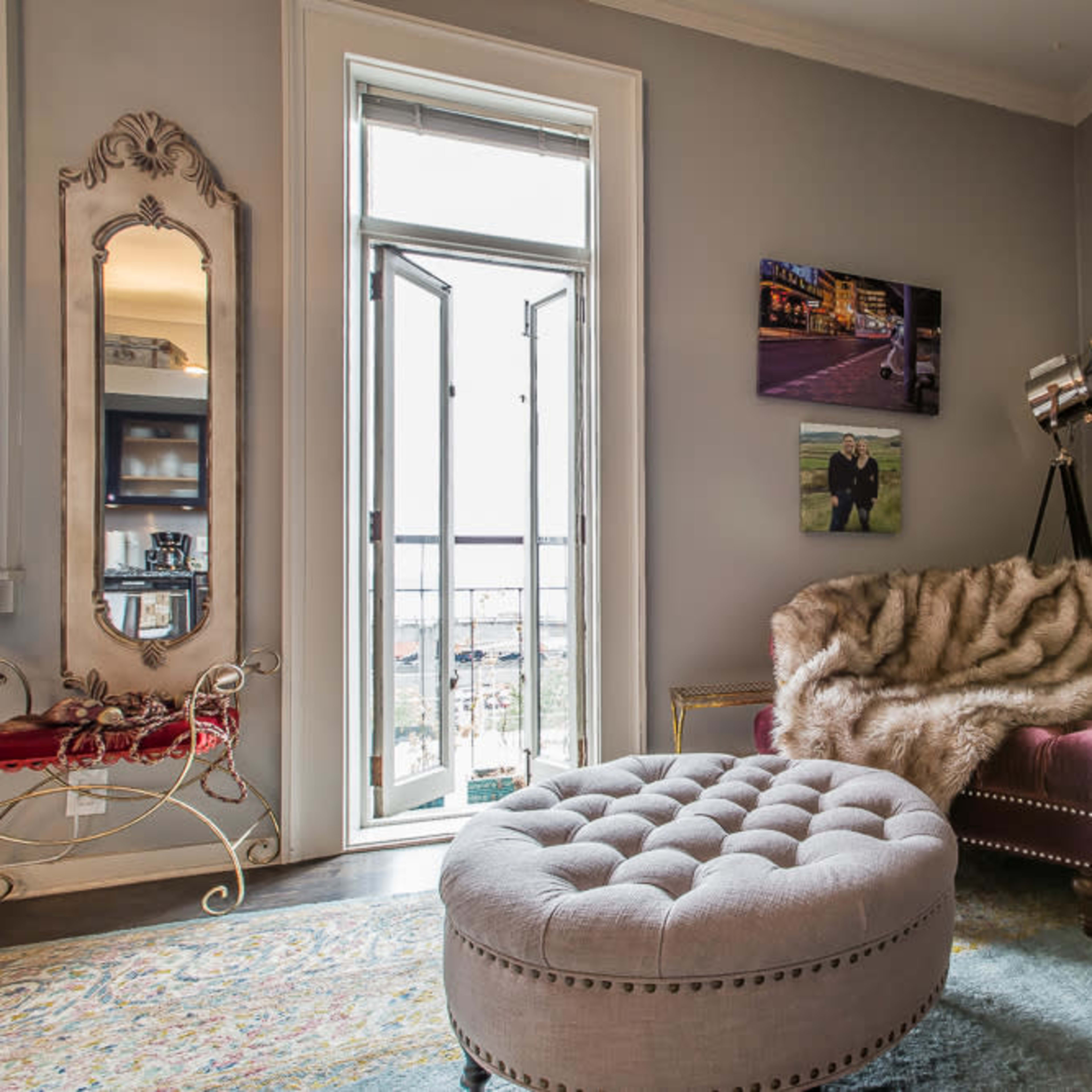 A cozy living room with a purple tufted sofa, a circular upholstered ottoman, and a balcony door allowing natural light to enter.