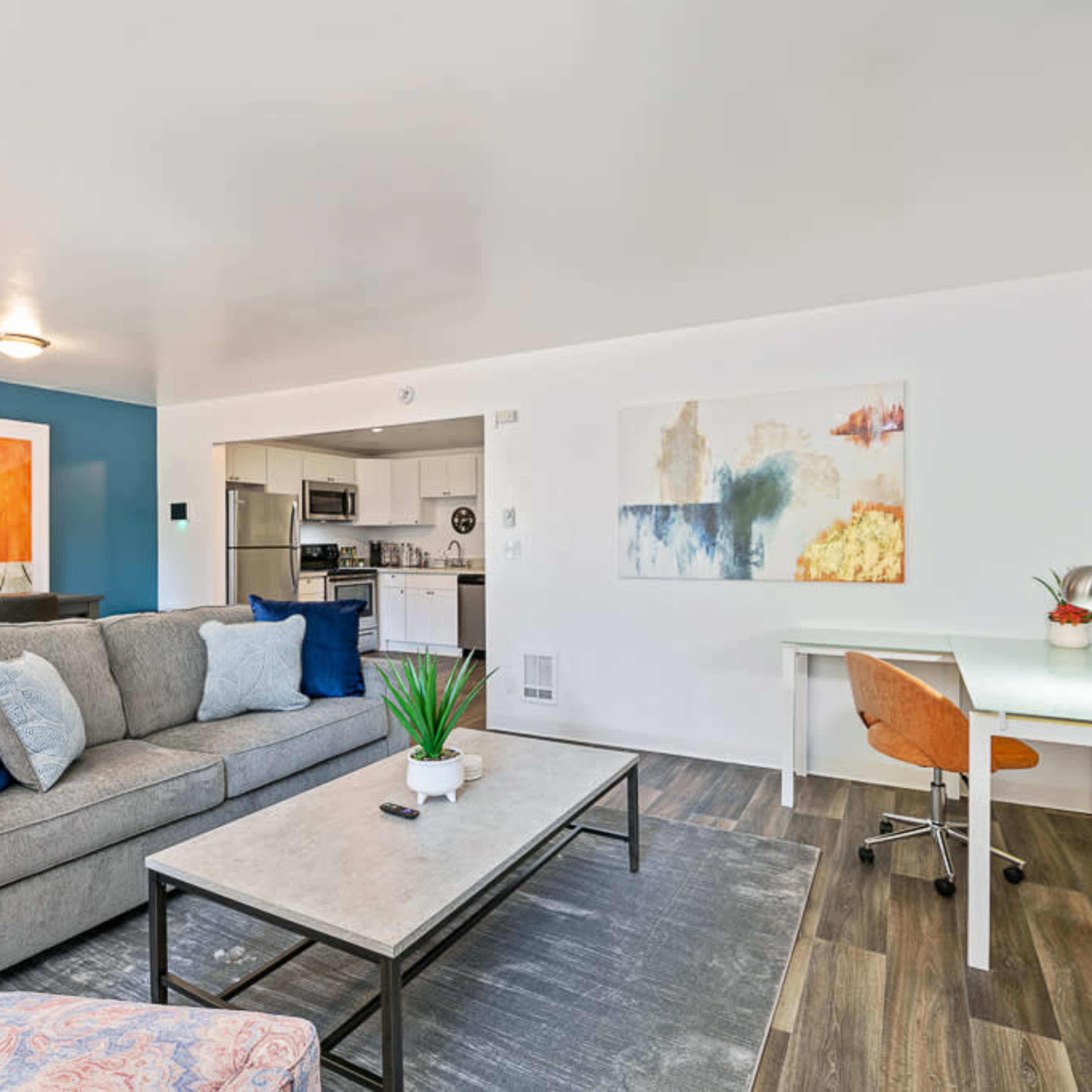 A modern living room with a gray couch, a coffee table, and a desk, featuring colorful artwork on the walls and a view into a kitchen area.