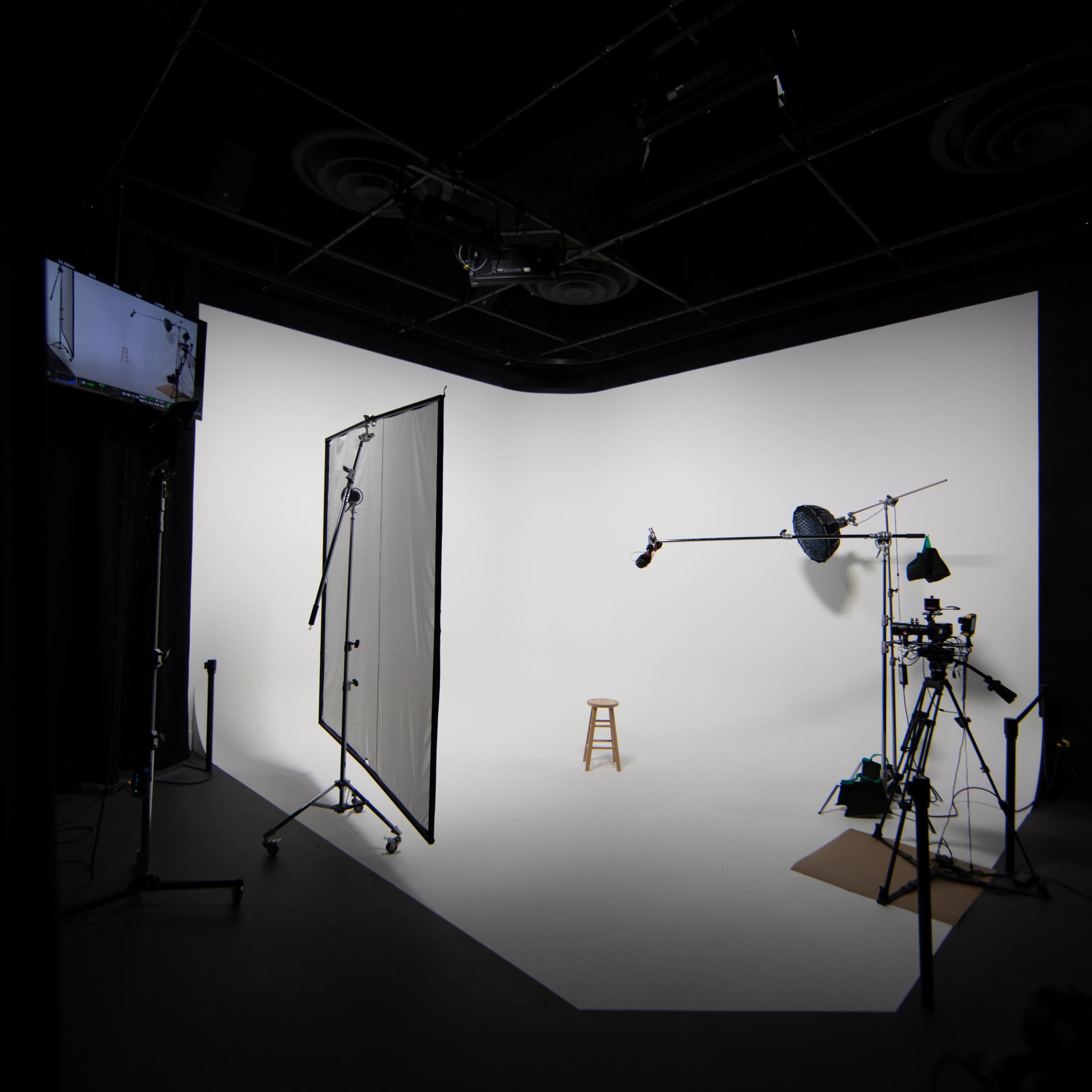 A photography studio featuring a white backdrop, a stool in the center, and various lighting and camera equipment arranged around the space.