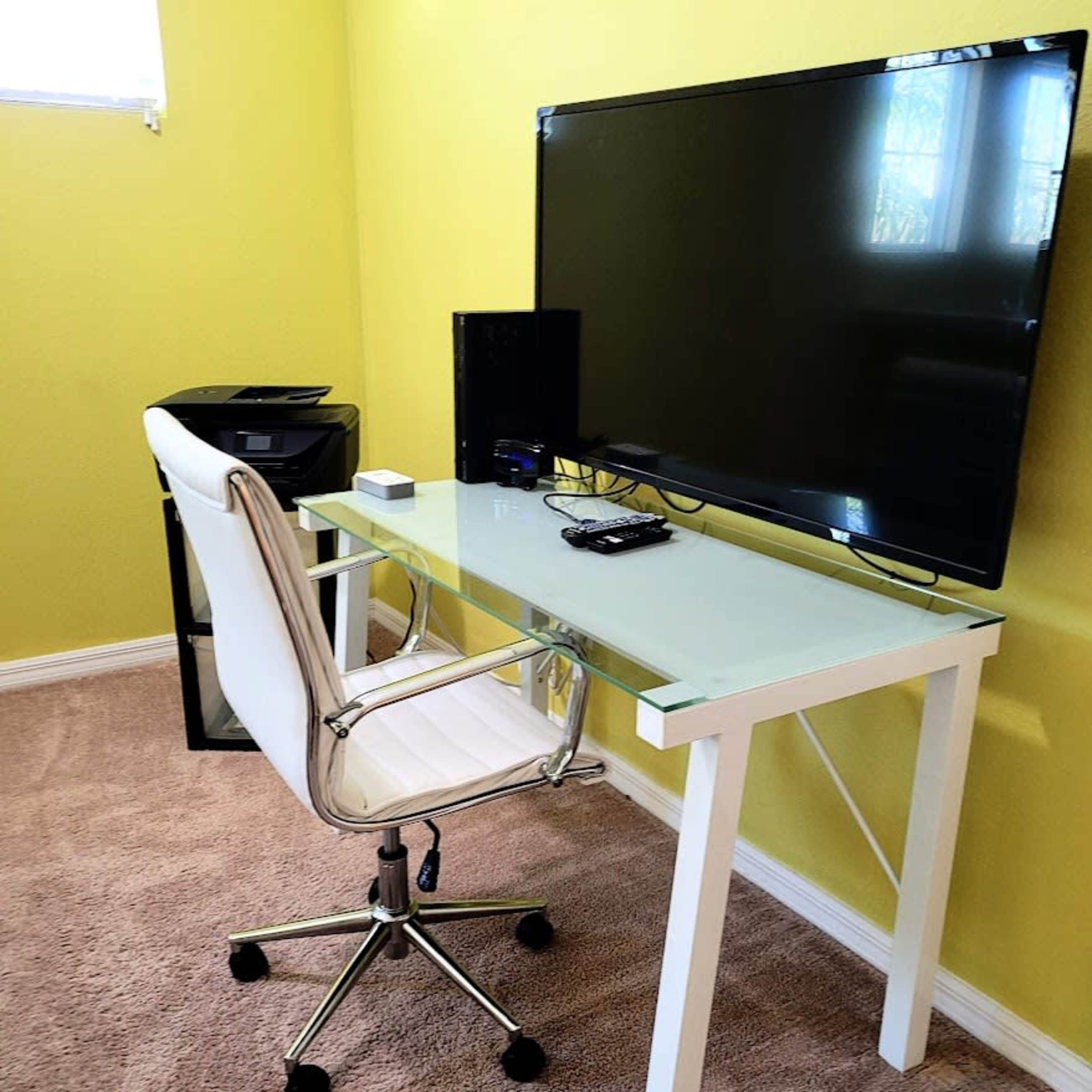 A modern desk setup featuring a glass-top desk, a white swivel chair, a large TV, and a small printer in a room with yellow walls.